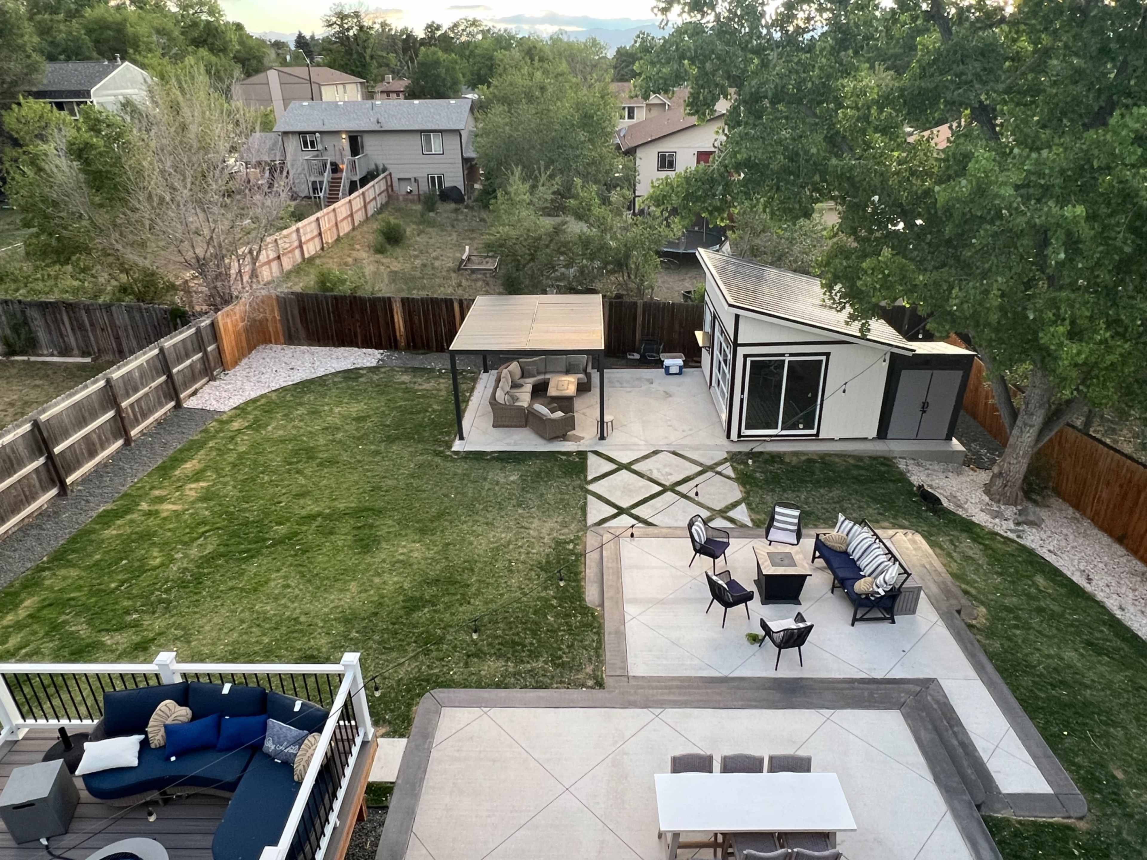 The image shows a spacious backyard with a grass area, a patio featuring outdoor furniture, and a modern structure in the corner.