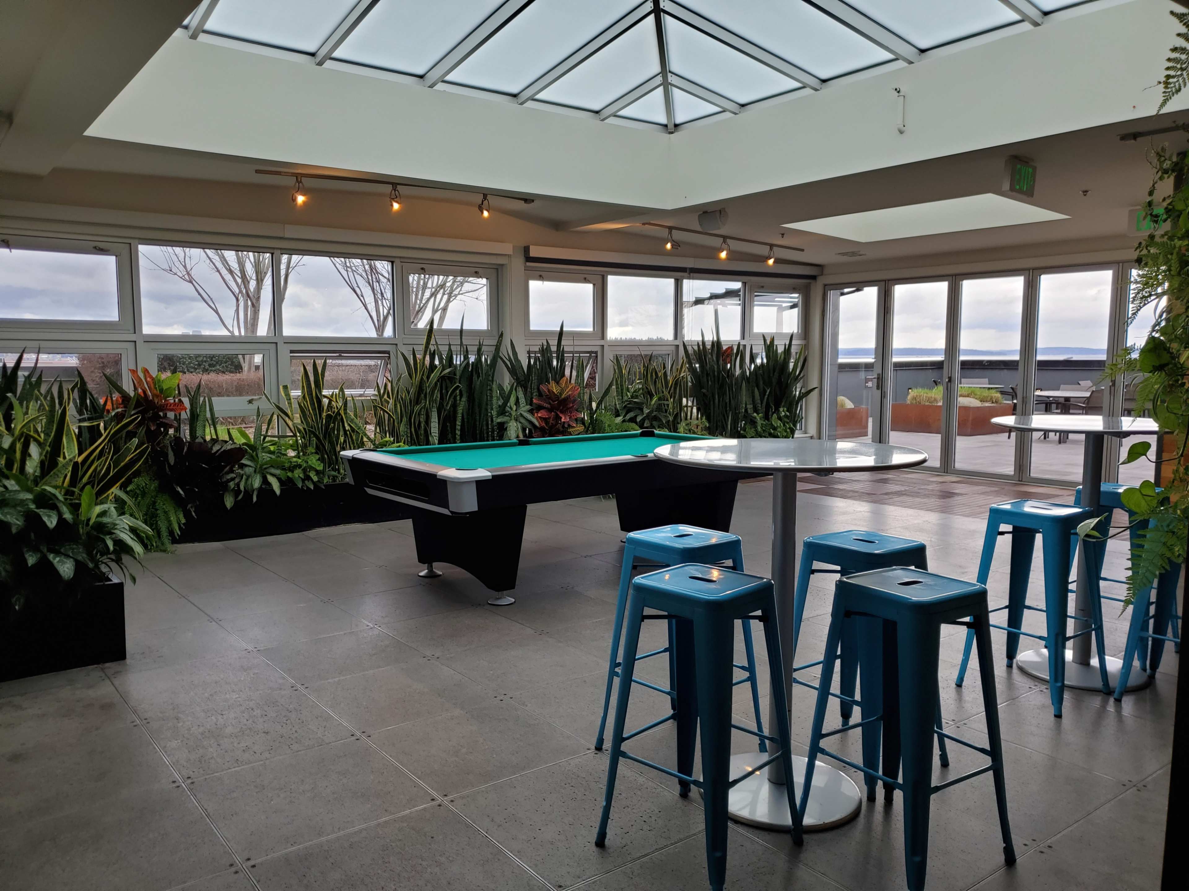 The spacious indoor area features a pool table, tall blue stools, and large windows with views of plants and a body of water beyond.