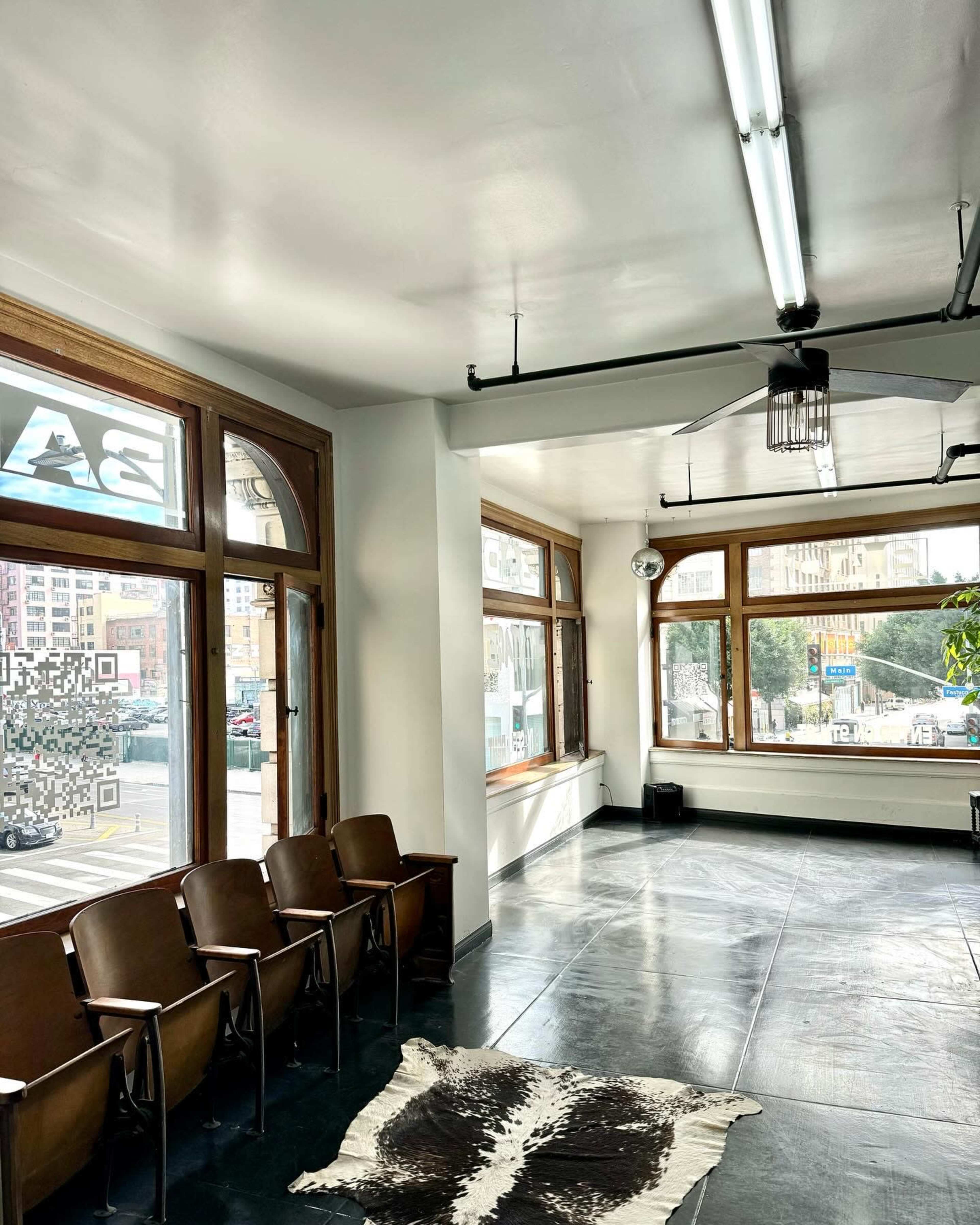 The image shows a minimalist interior of a room with large windows, wooden chairs arranged in a row, and a cowhide rug on the floor.
