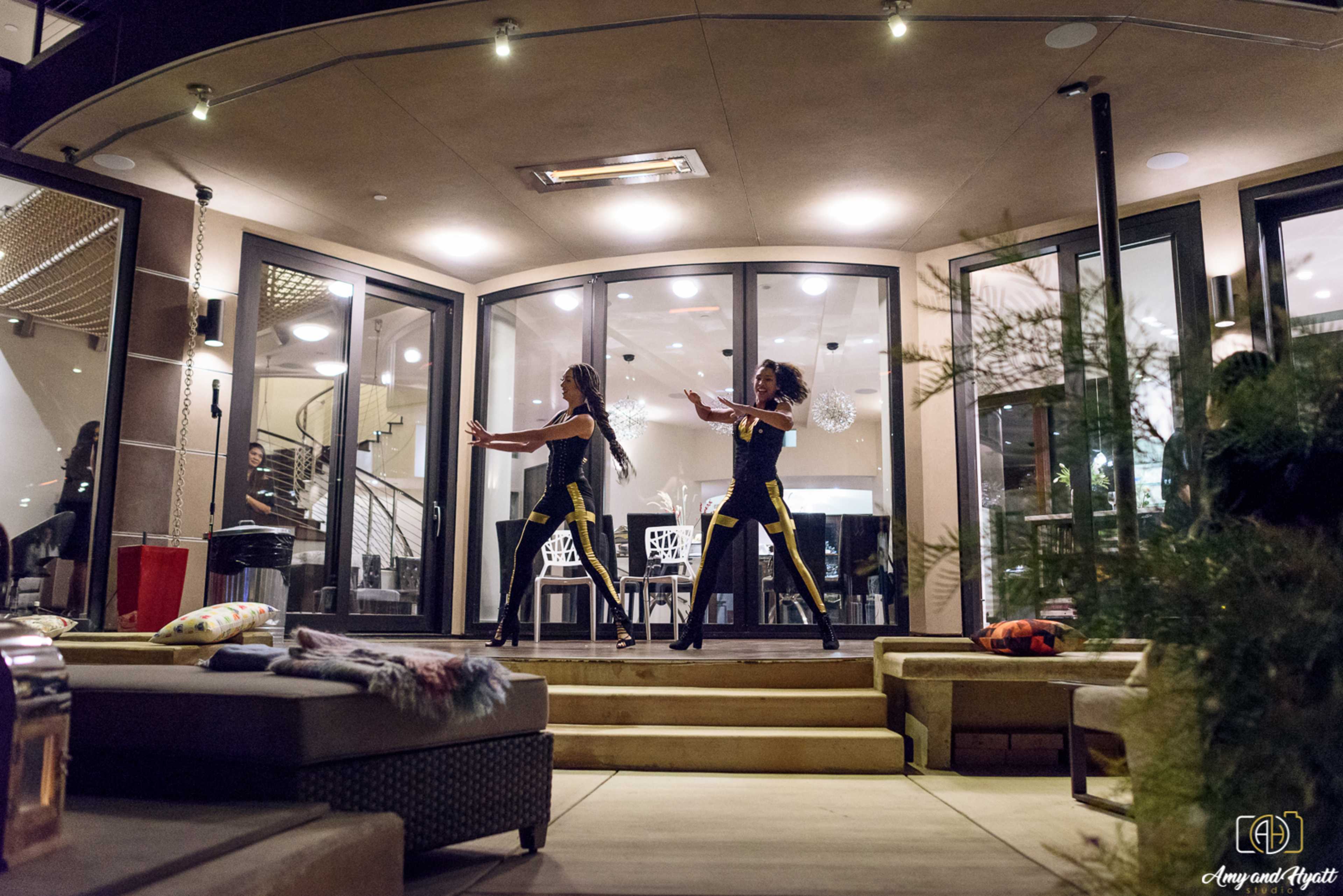 Two dancers perform on a patio in front of a large glass door, showcasing a contemporary style against an interior lit background.