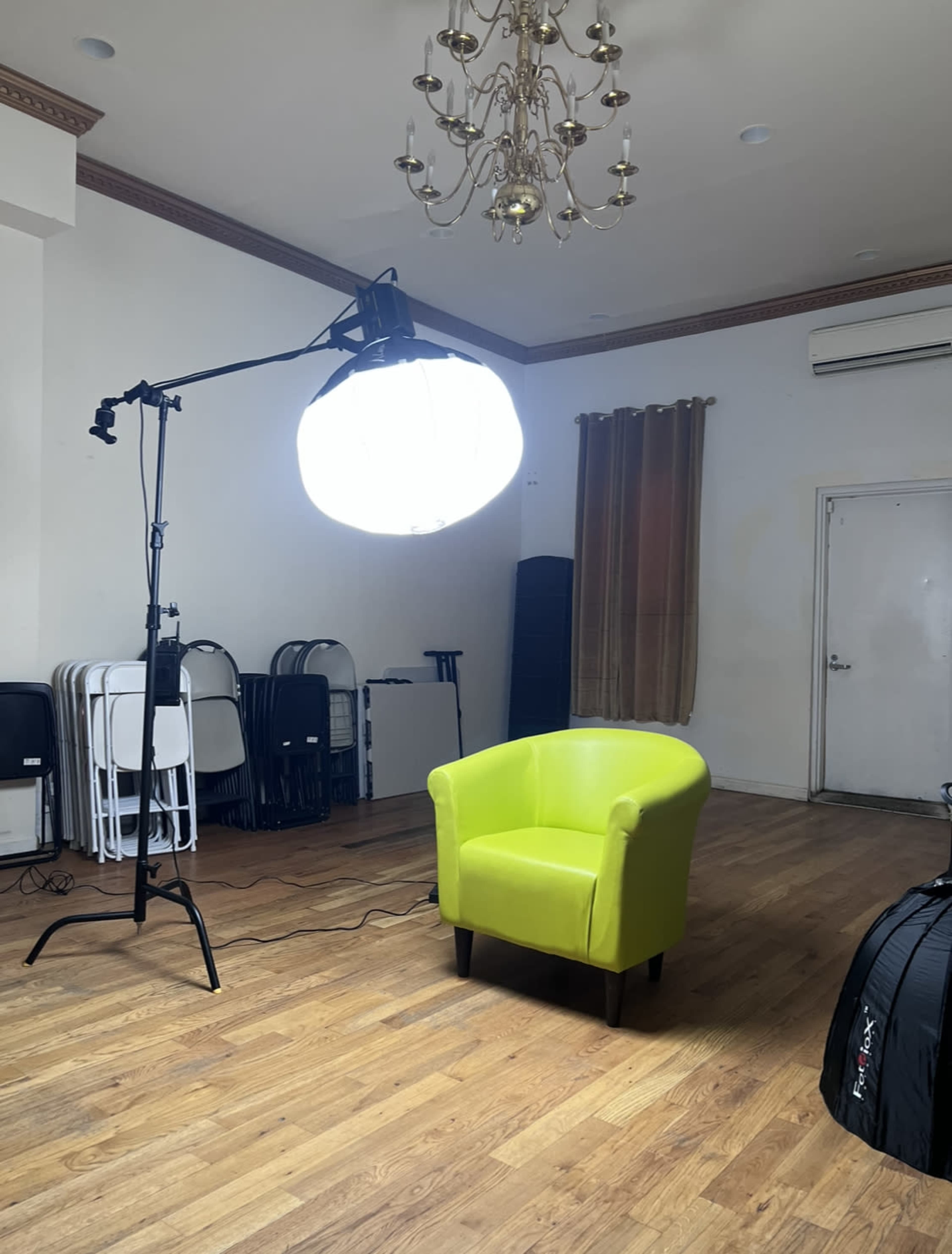 A bright green chair placed in an empty room illuminated by a large softbox light, with folding chairs stacked in the background.