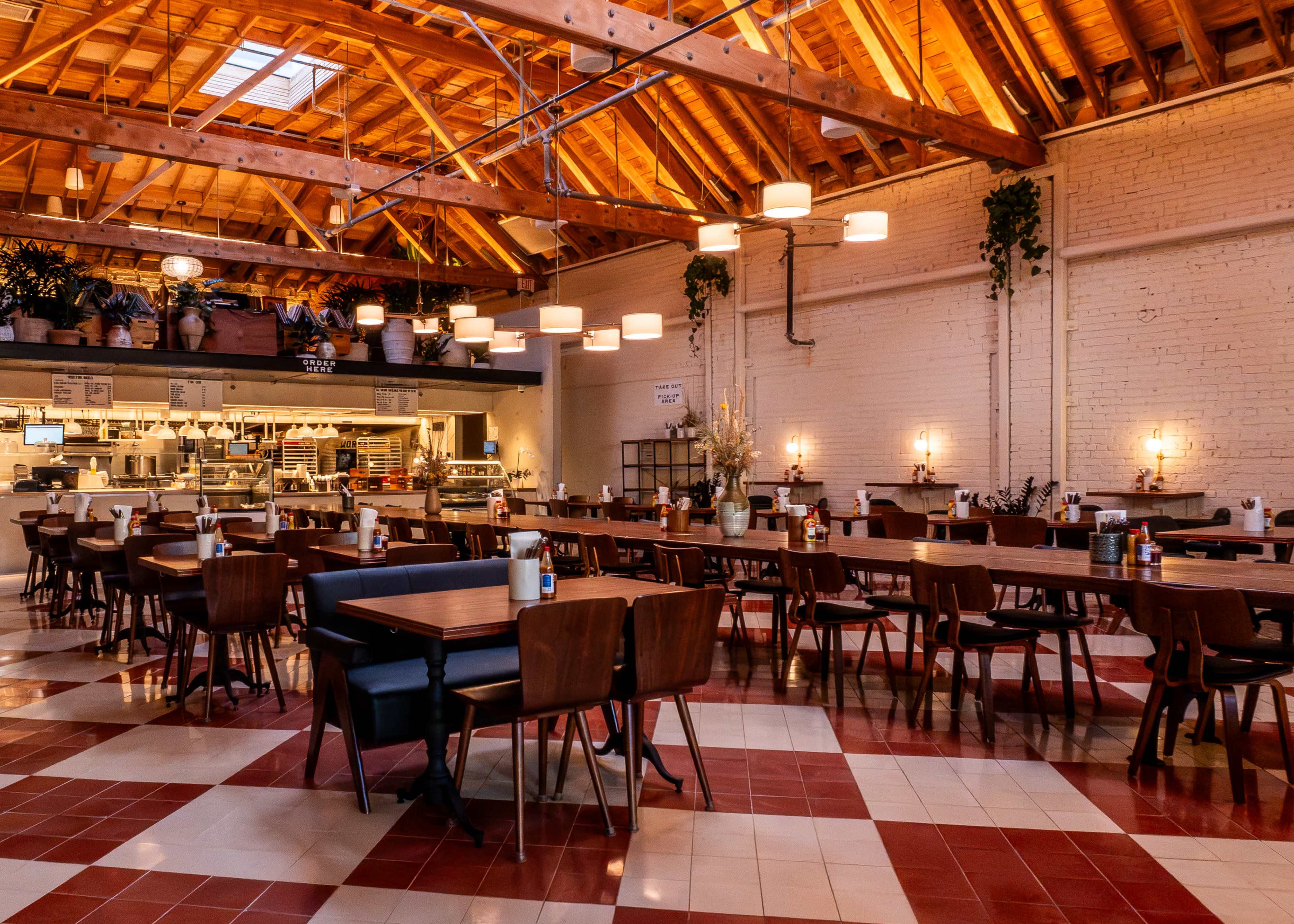 The image shows a spacious restaurant with wooden tables and chairs, featuring a high ceiling with exposed beams and multiple light fixtures.