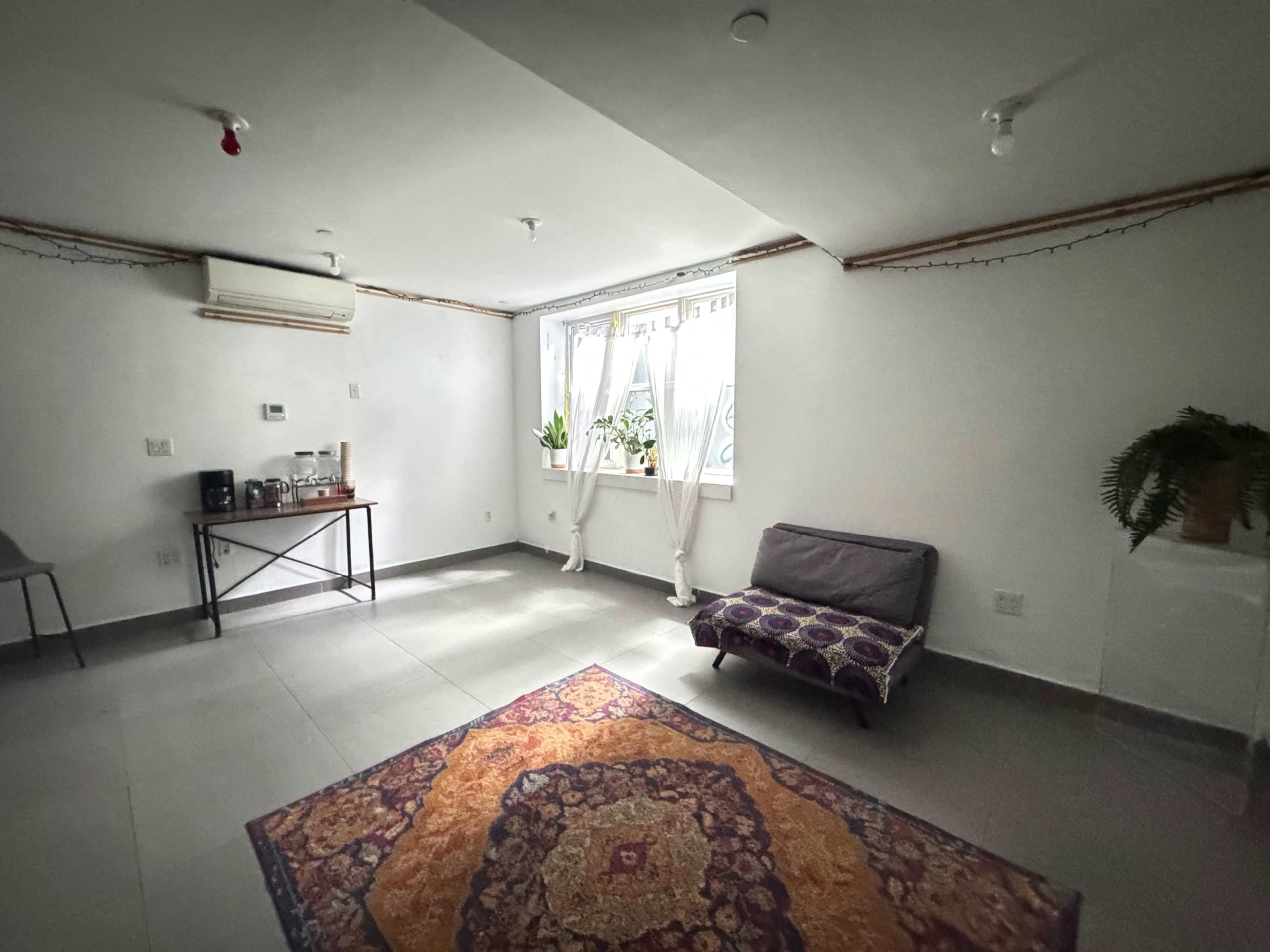 The image shows a spacious, minimally furnished room with a patterned rug, a foldable sofa, a small table with coffee supplies, and windows with sheer curtains.