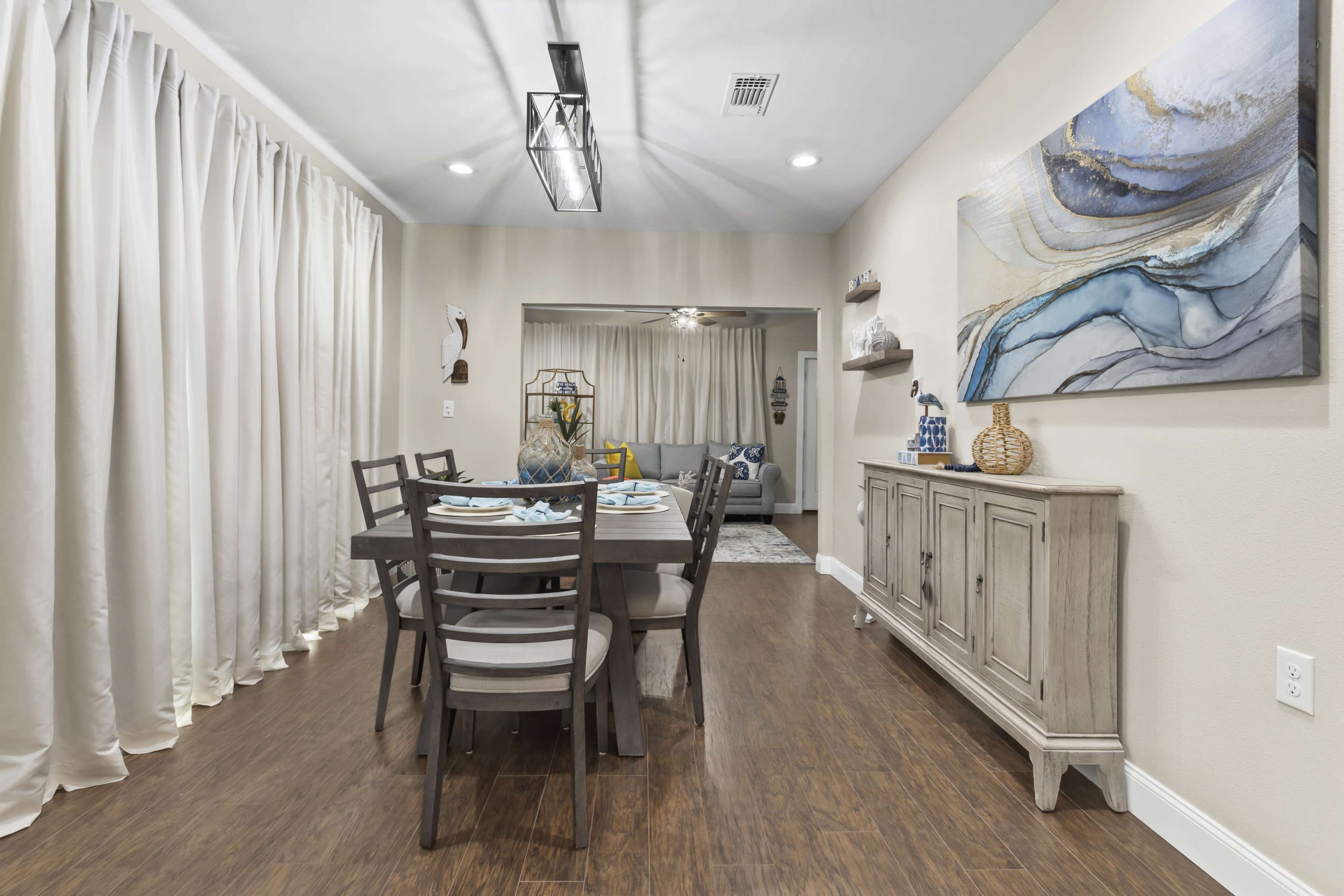 A dining area with a rectangular table set for a meal, accompanied by a sideboard and a wall adorned with abstract art and soft curtain drapes.