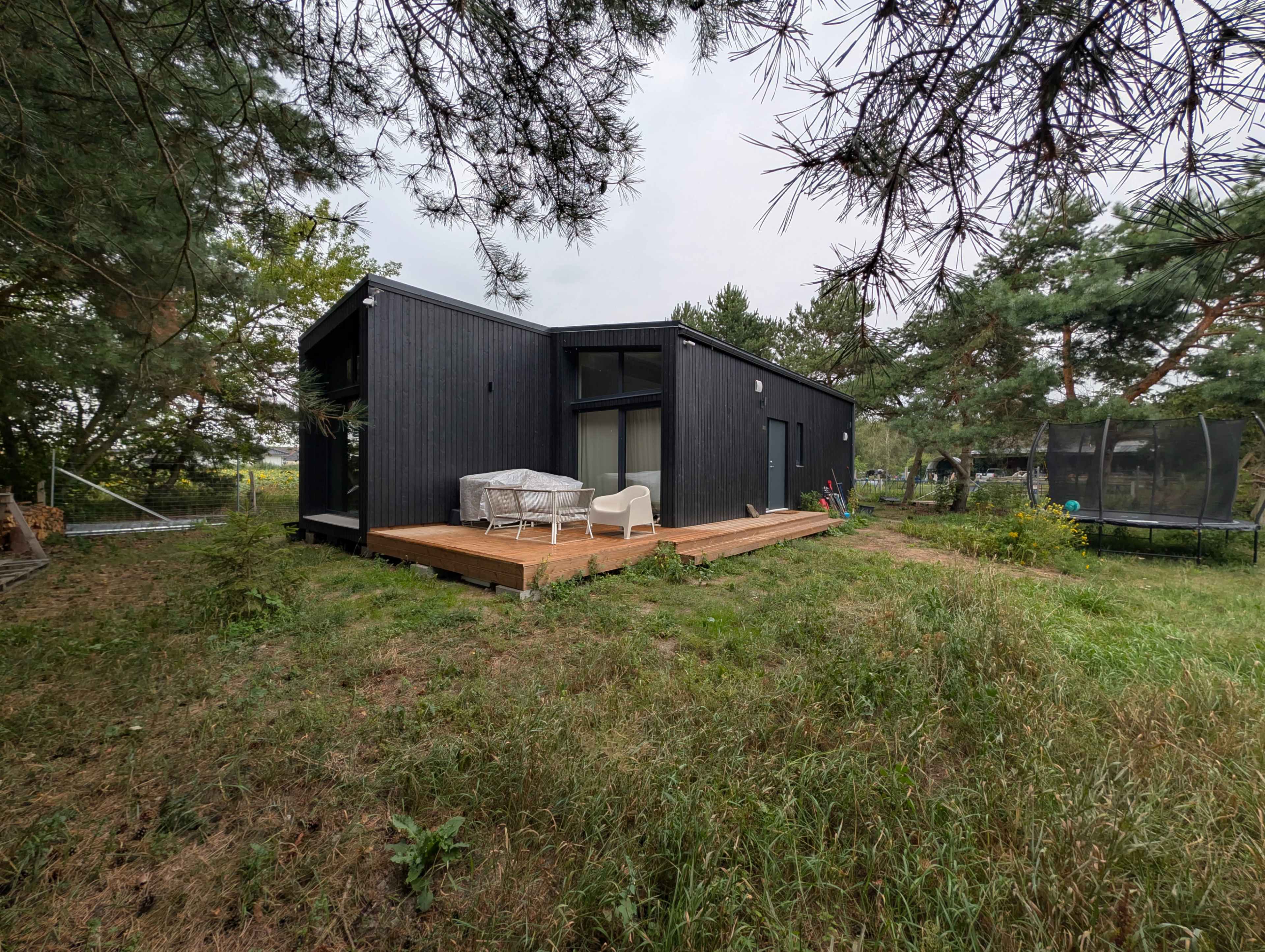 The image shows a modern black wooden house with a wooden deck surrounded by grass and trees.