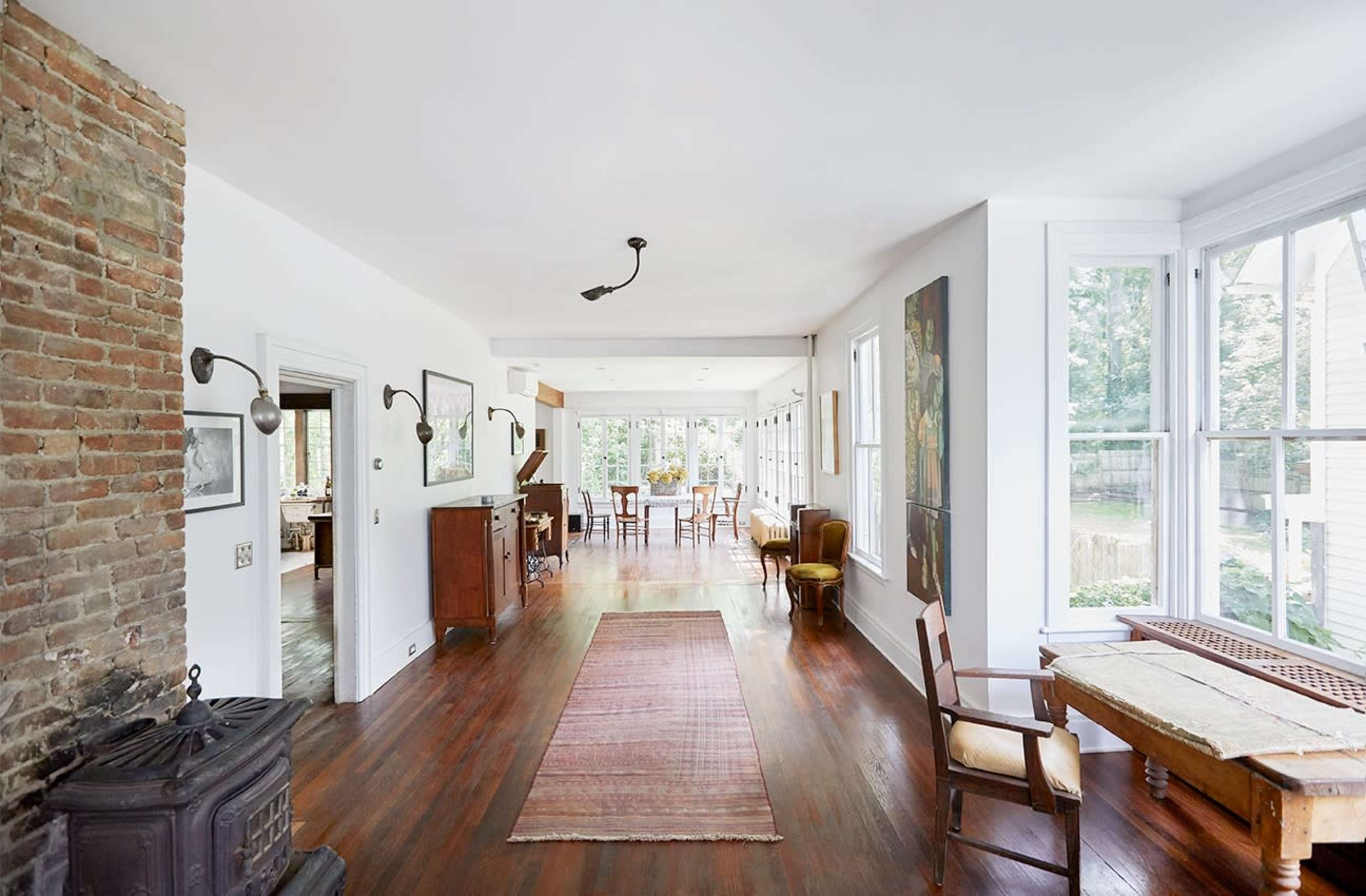 A spacious, bright interior of a home with wooden flooring, a brick wall, and large windows overlooking a garden area.