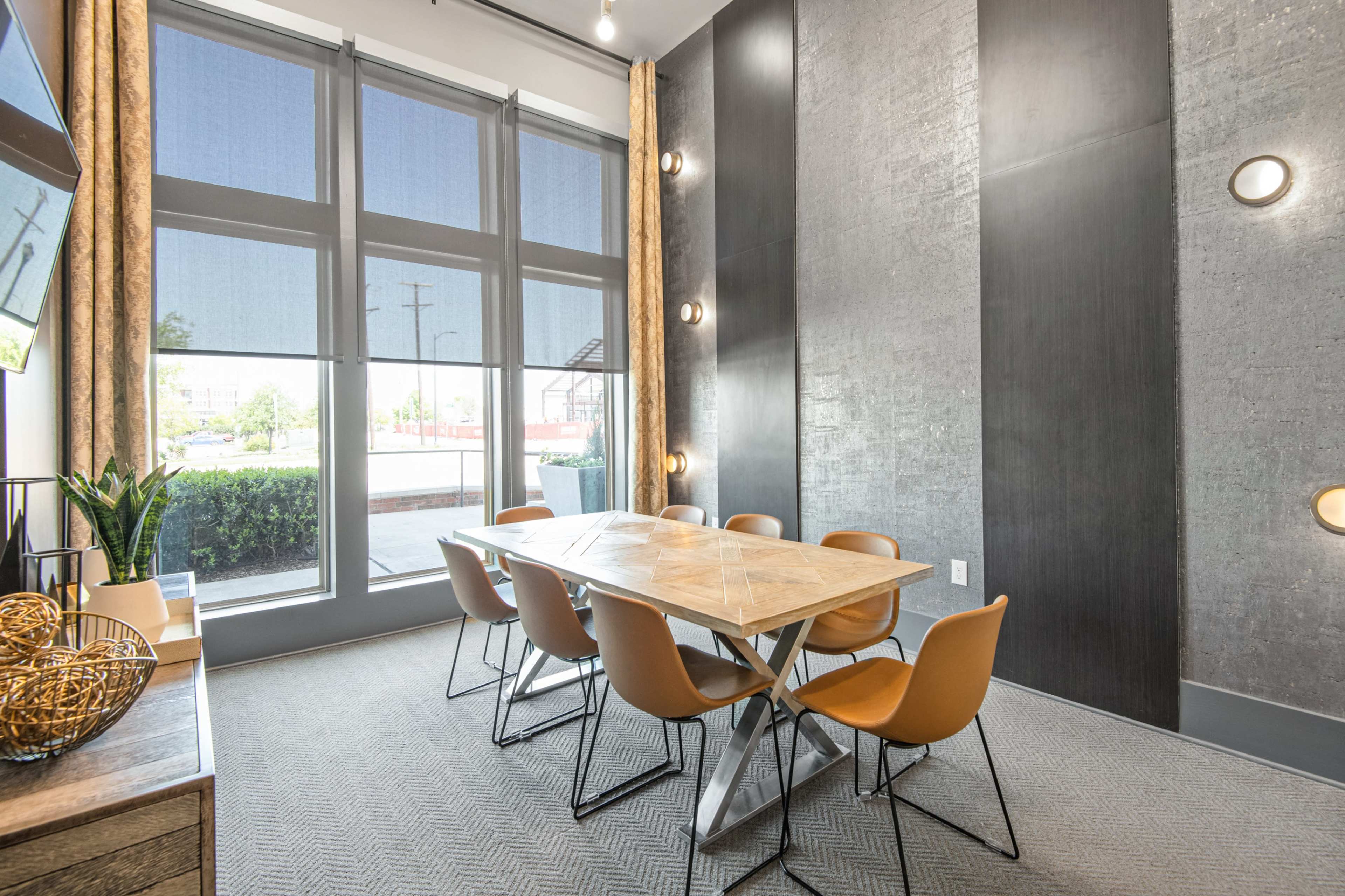 A conference room features a long wooden table surrounded by six orange chairs, with large windows providing natural light.