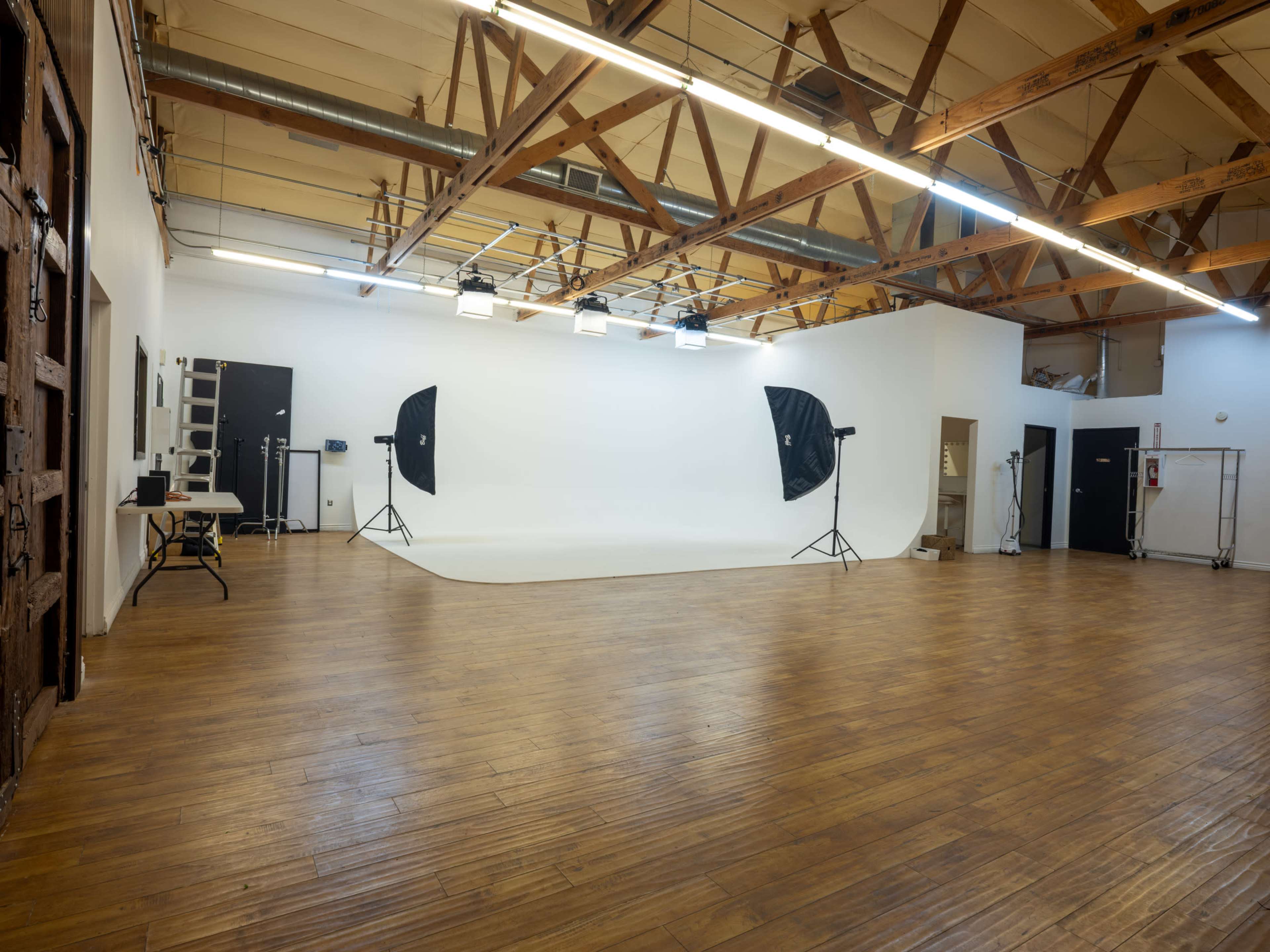 The image shows a spacious photography studio with wooden floors, a white backdrop, and two black softbox lights set up on stands.