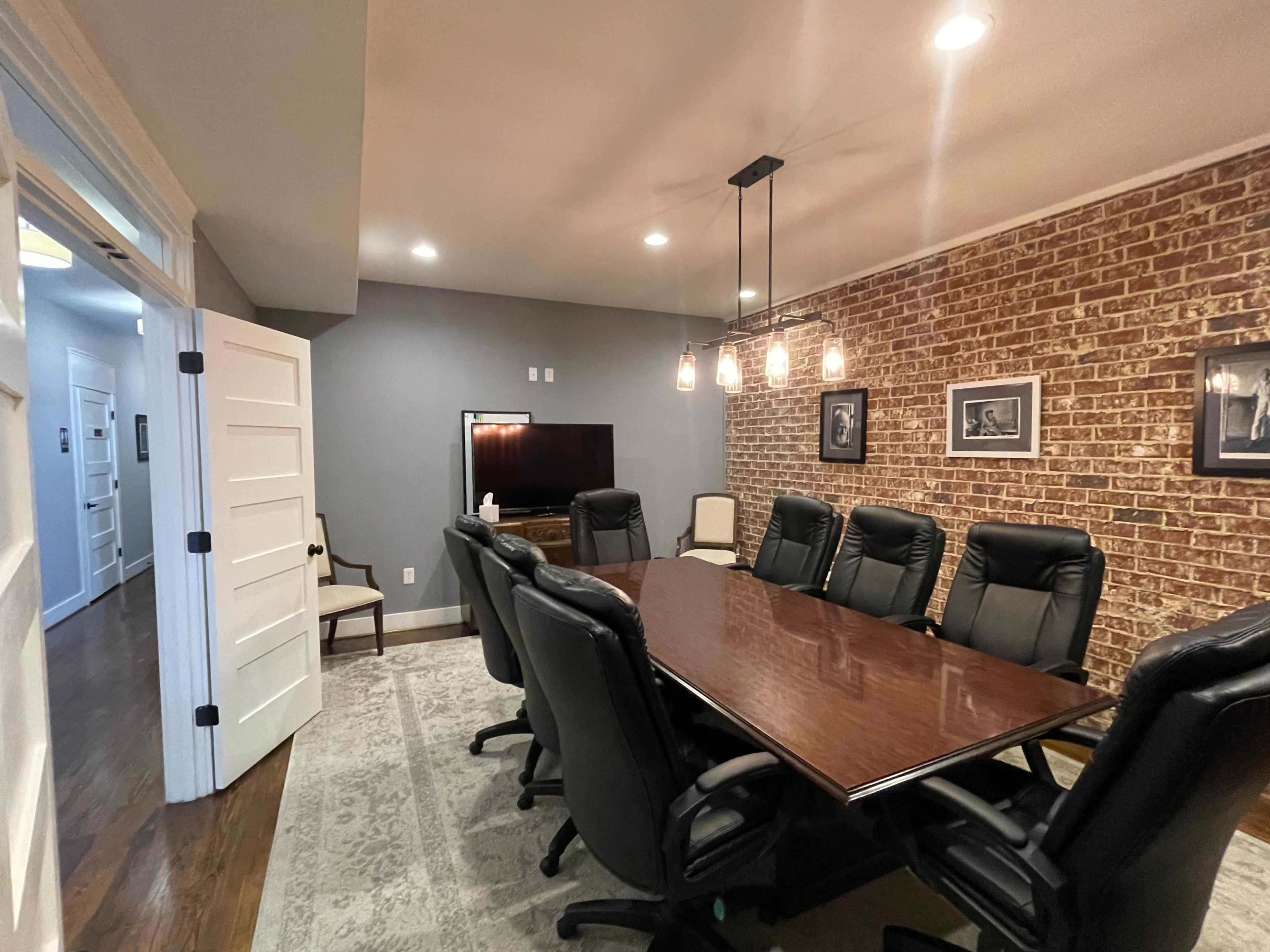 A conference room with a long wooden table surrounded by black leather chairs, a television mounted on the wall, and exposed brick accents.