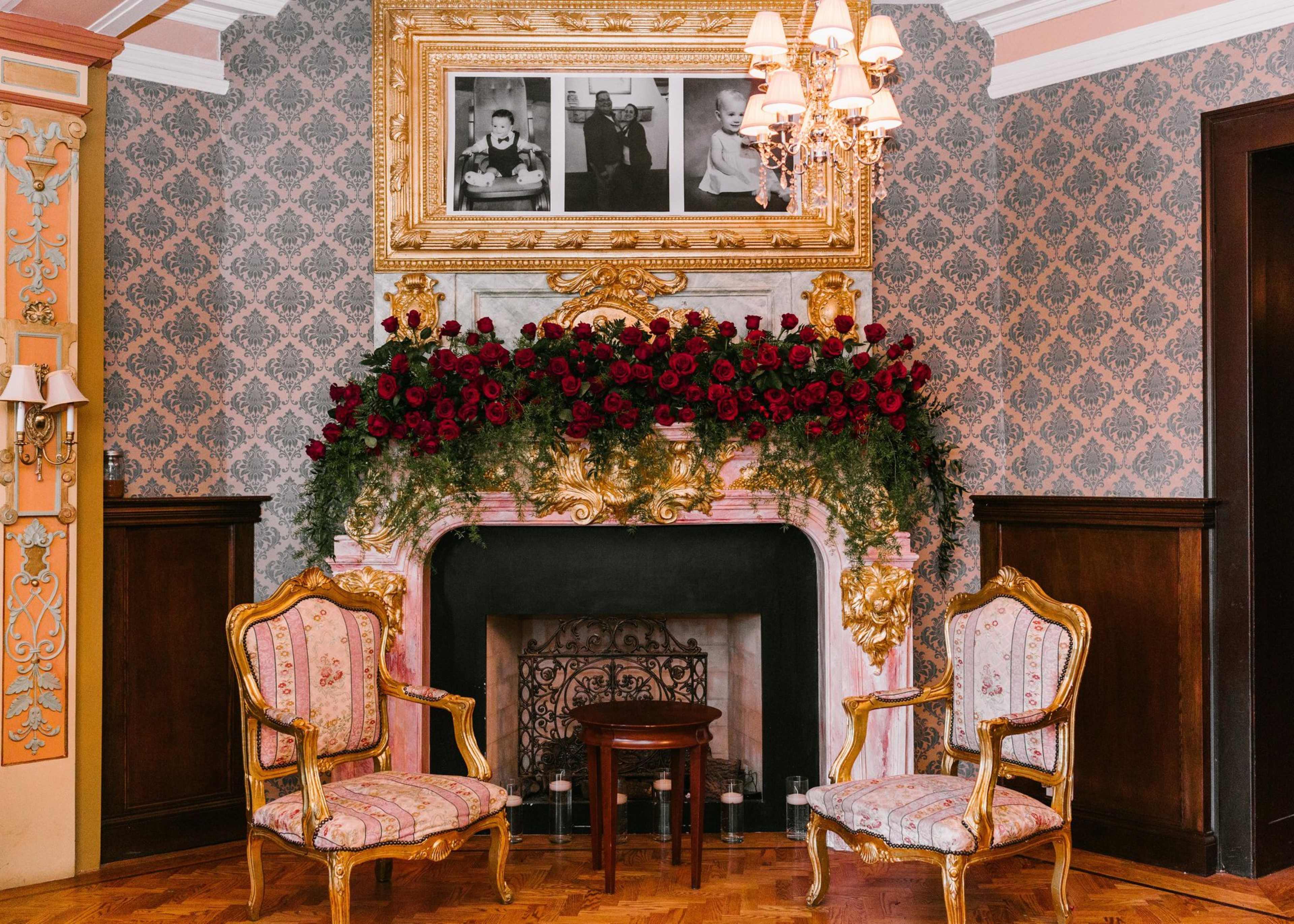 An elegantly decorated room with a floral arrangement of red roses on a marble fireplace, framed photos above, and two ornate gold chairs facing a small table.