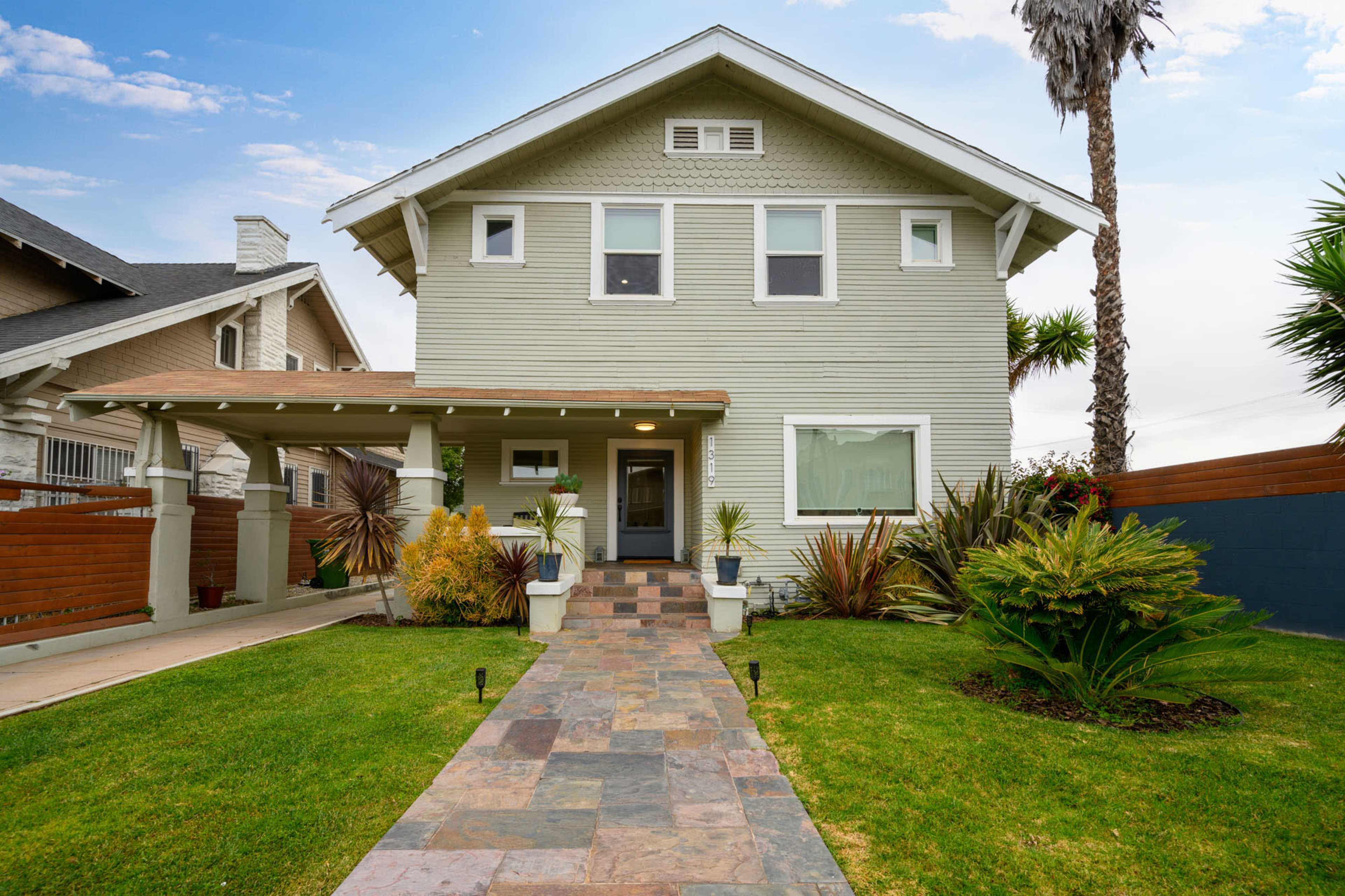 A two-story house with a light green exterior and a landscaped front yard featuring a stone pathway and various plants.