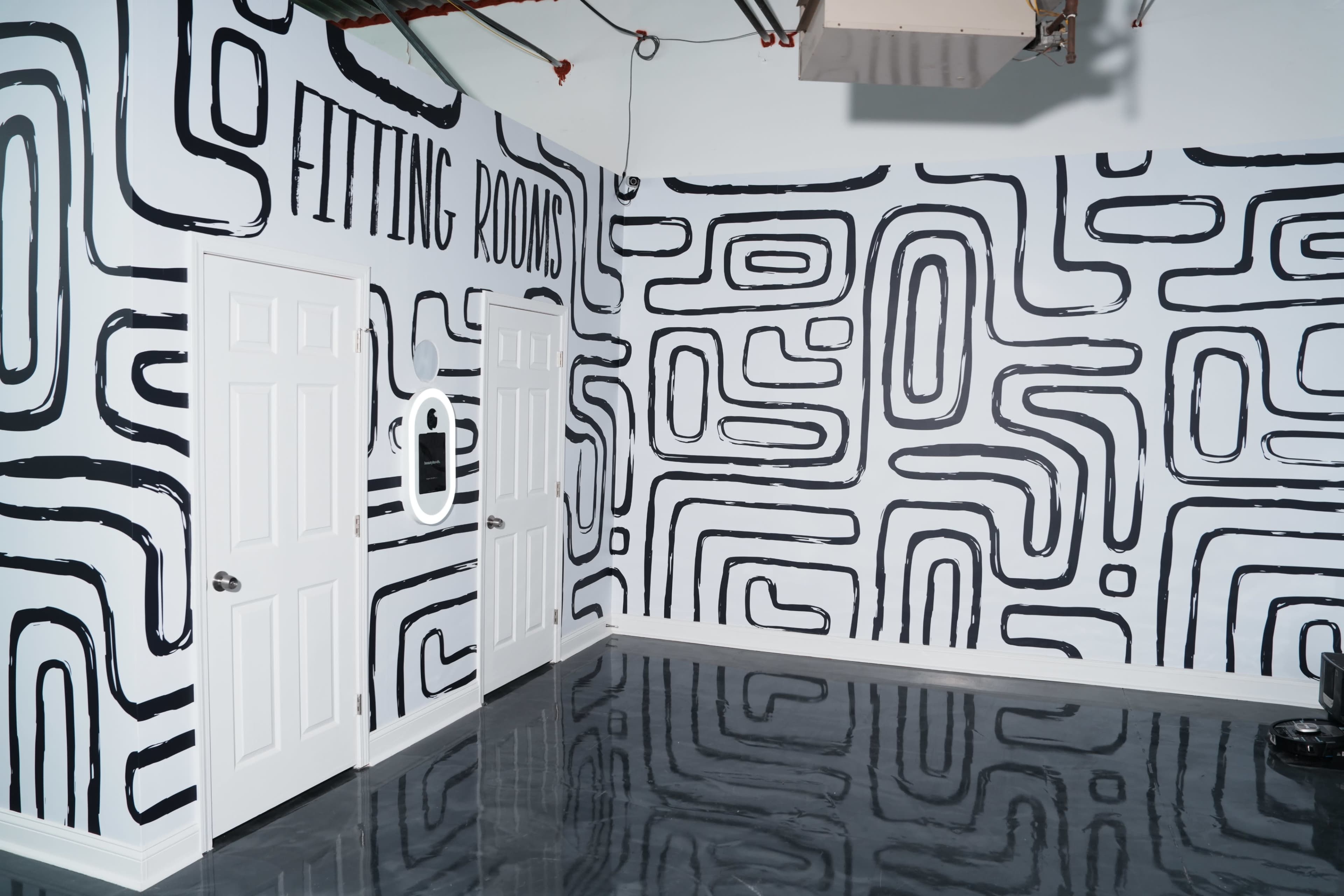 The image shows a room with black and white patterned walls featuring the words "FITTING ROOMS," along with two doors and a mirror on one wall.