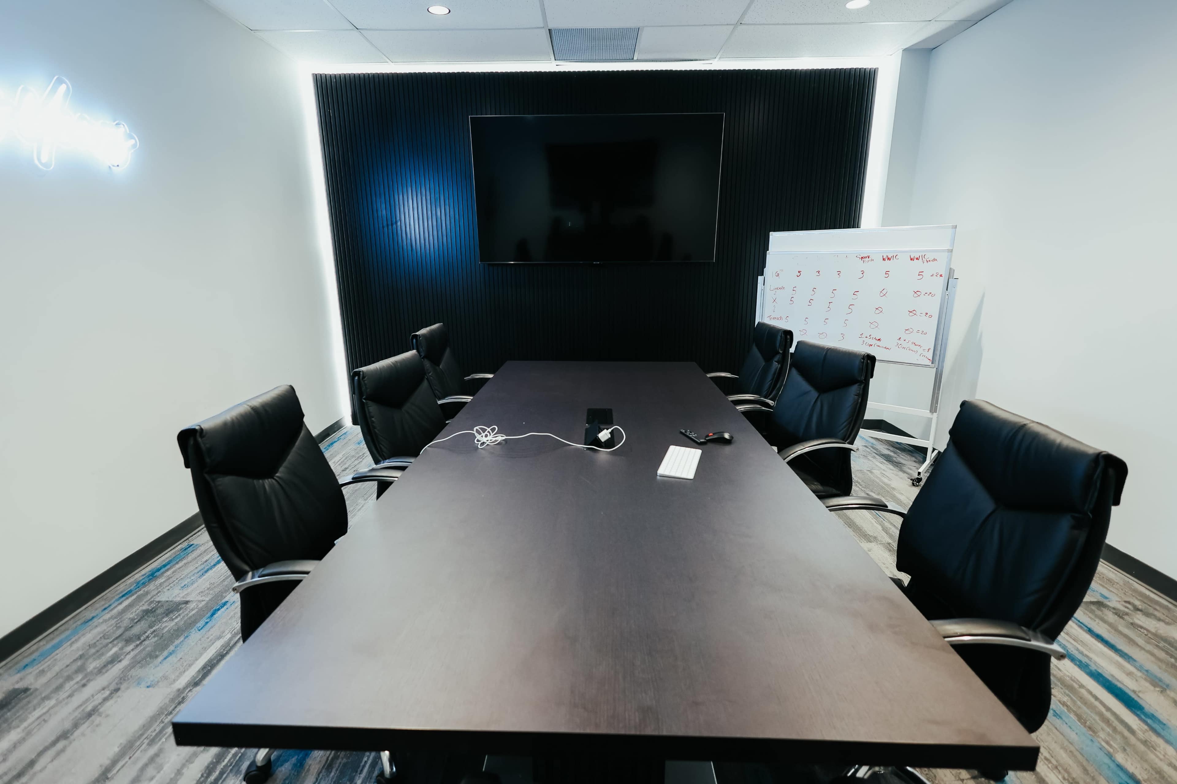 A conference room features a long table surrounded by black chairs, with a large screen mounted on the wall and a whiteboard in the corner.