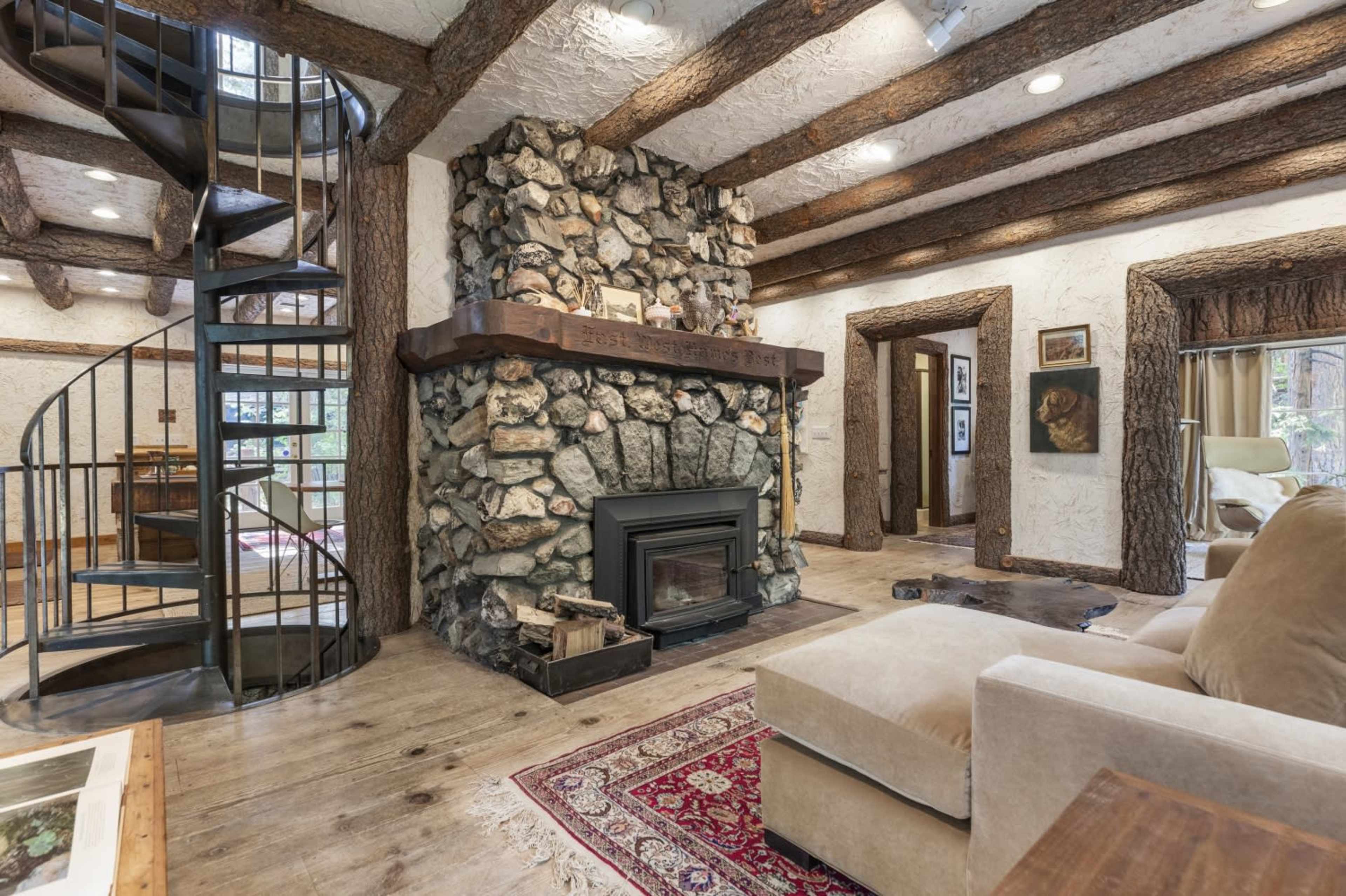 The image shows a rustic living room with a spiral staircase, a stone fireplace, and wood-beamed ceilings.