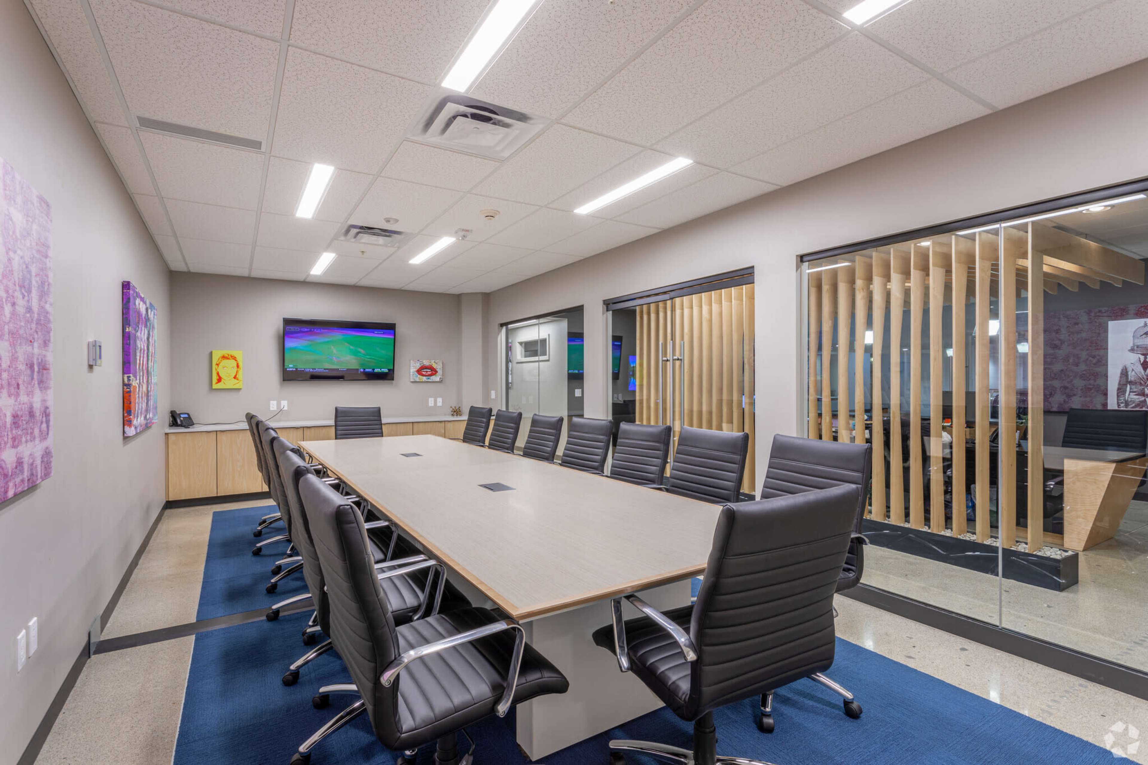 A modern conference room with a long rectangular table surrounded by black office chairs and a glass wall separating it from an adjacent room.