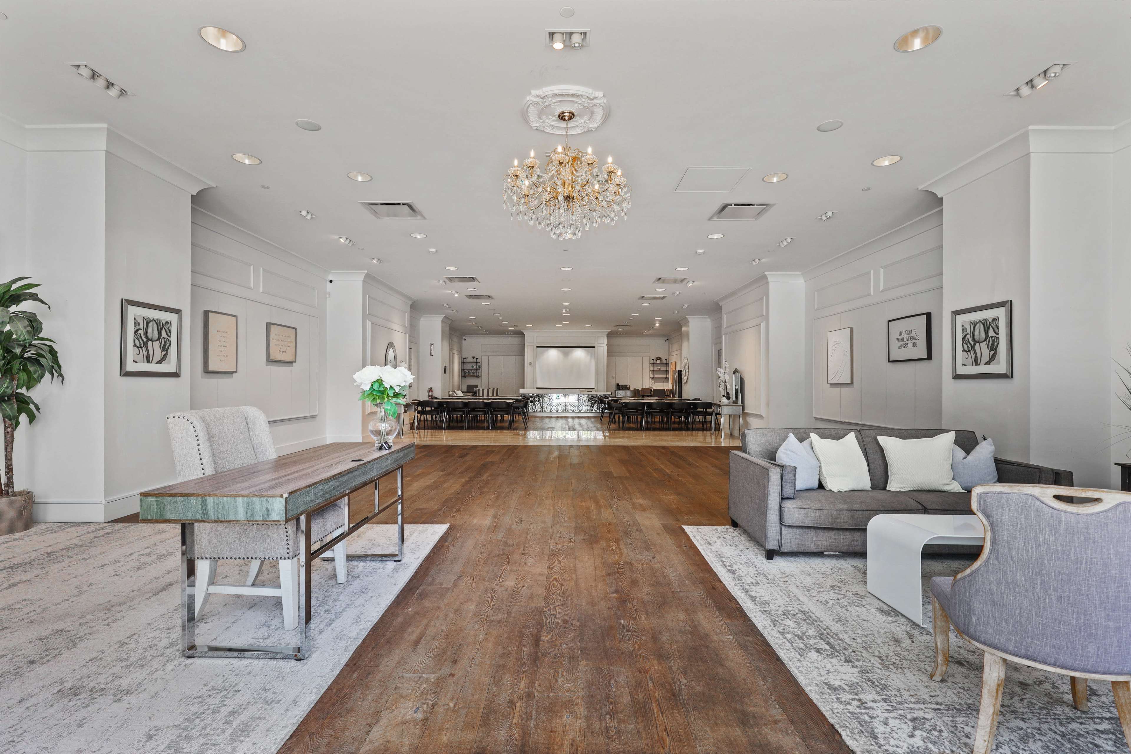 The image shows a spacious event venue featuring a wooden floor, a chandelier, and a seating area with a sofa and a table at one end.
