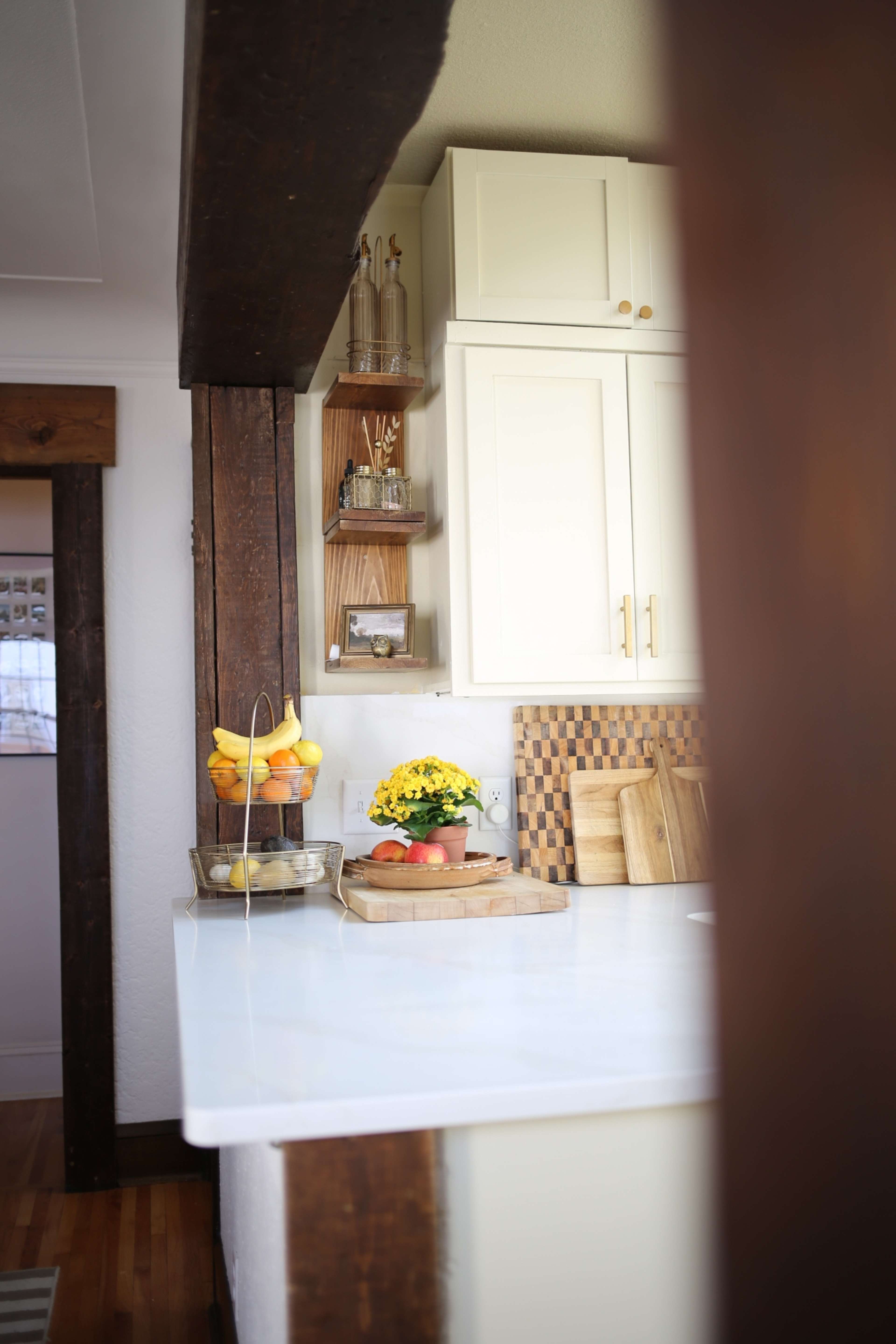 The image shows a kitchen with white cabinets, a wooden shelf holding decorative items, and a countertop featuring a fruit bowl and a potted plant.