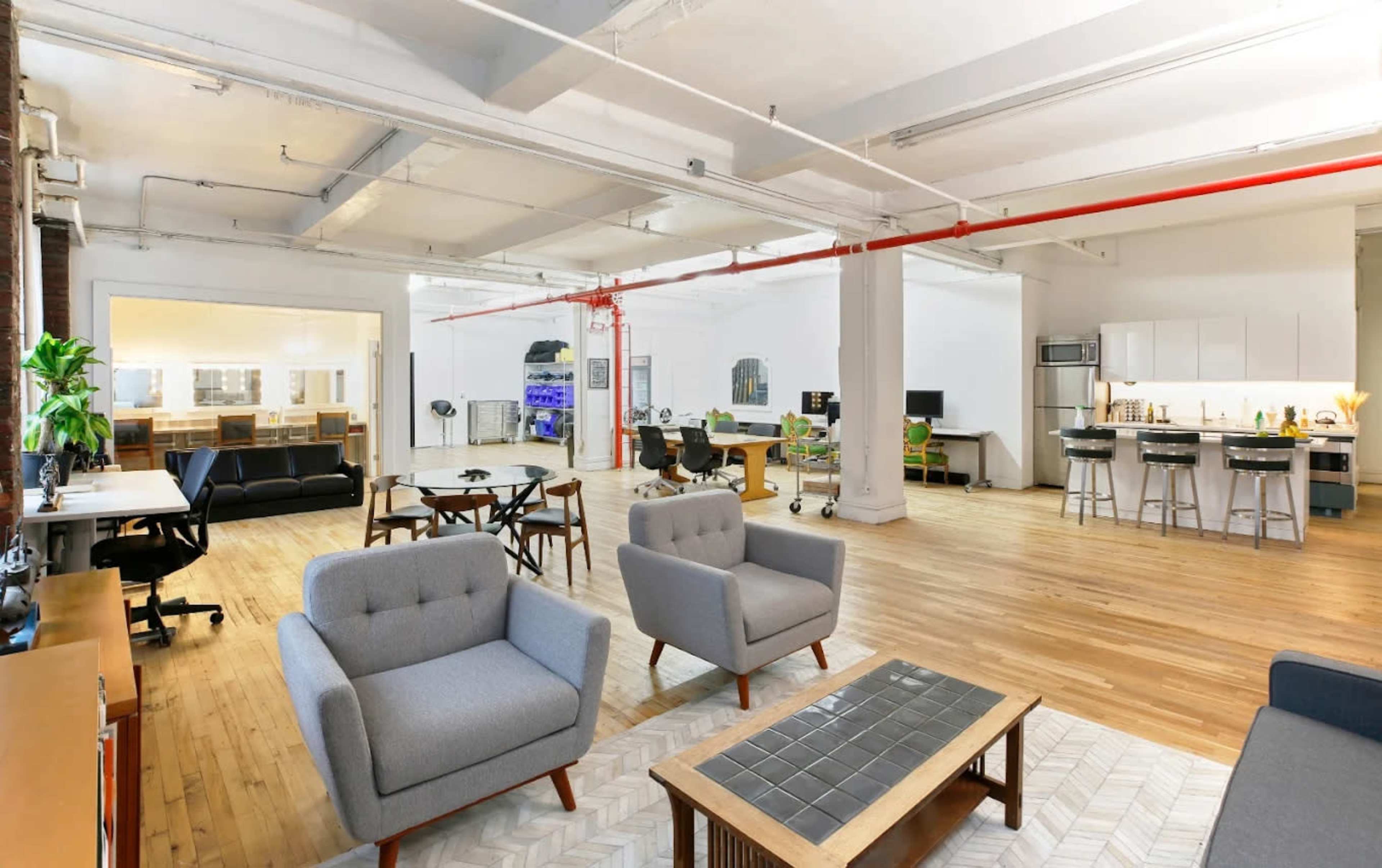 The image shows a spacious office interior featuring modern furniture, wooden flooring, and workstations along with a kitchen area.
