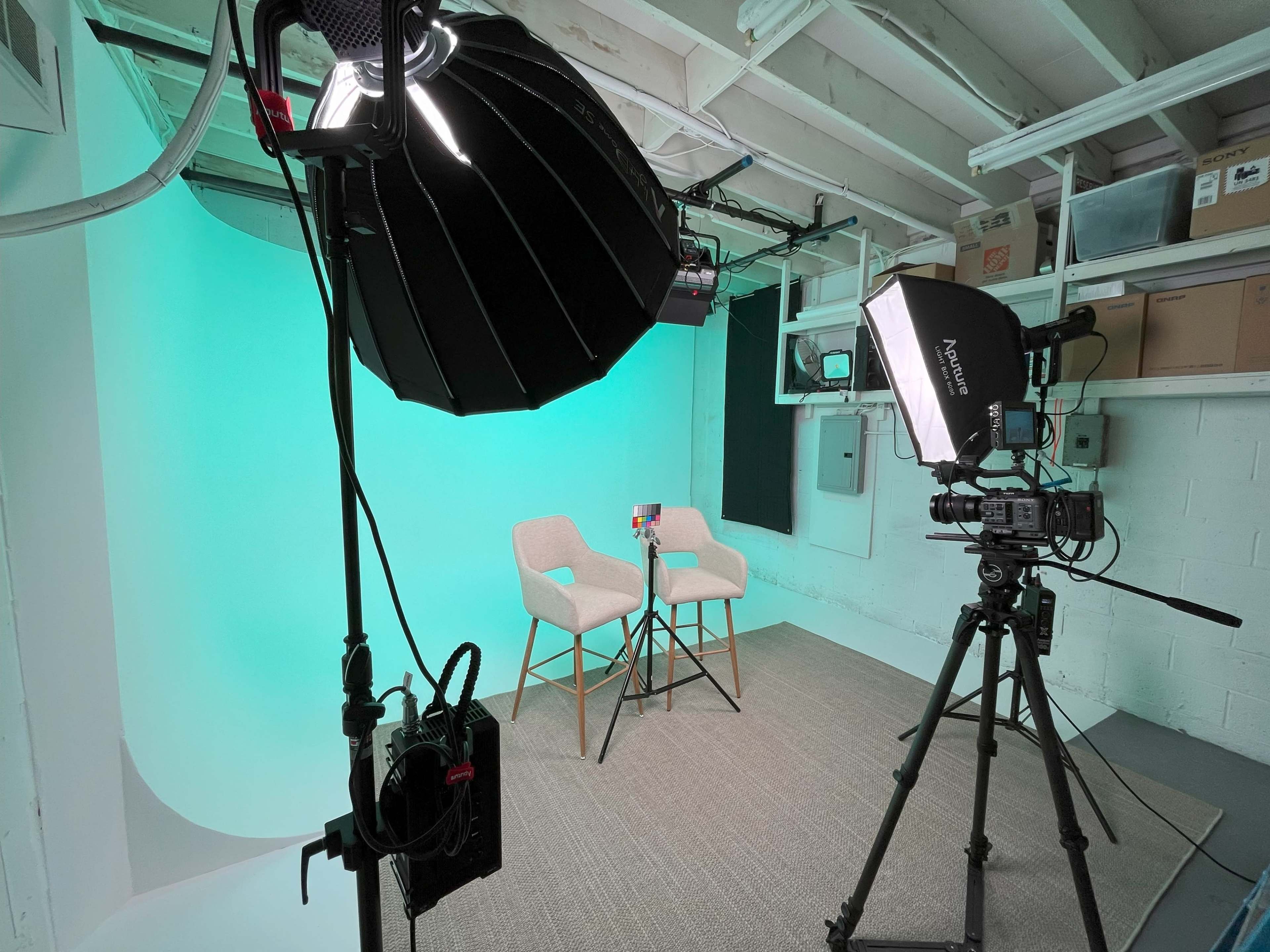A studio setup with two chairs facing a camera, surrounded by professional lighting and a green backdrop.