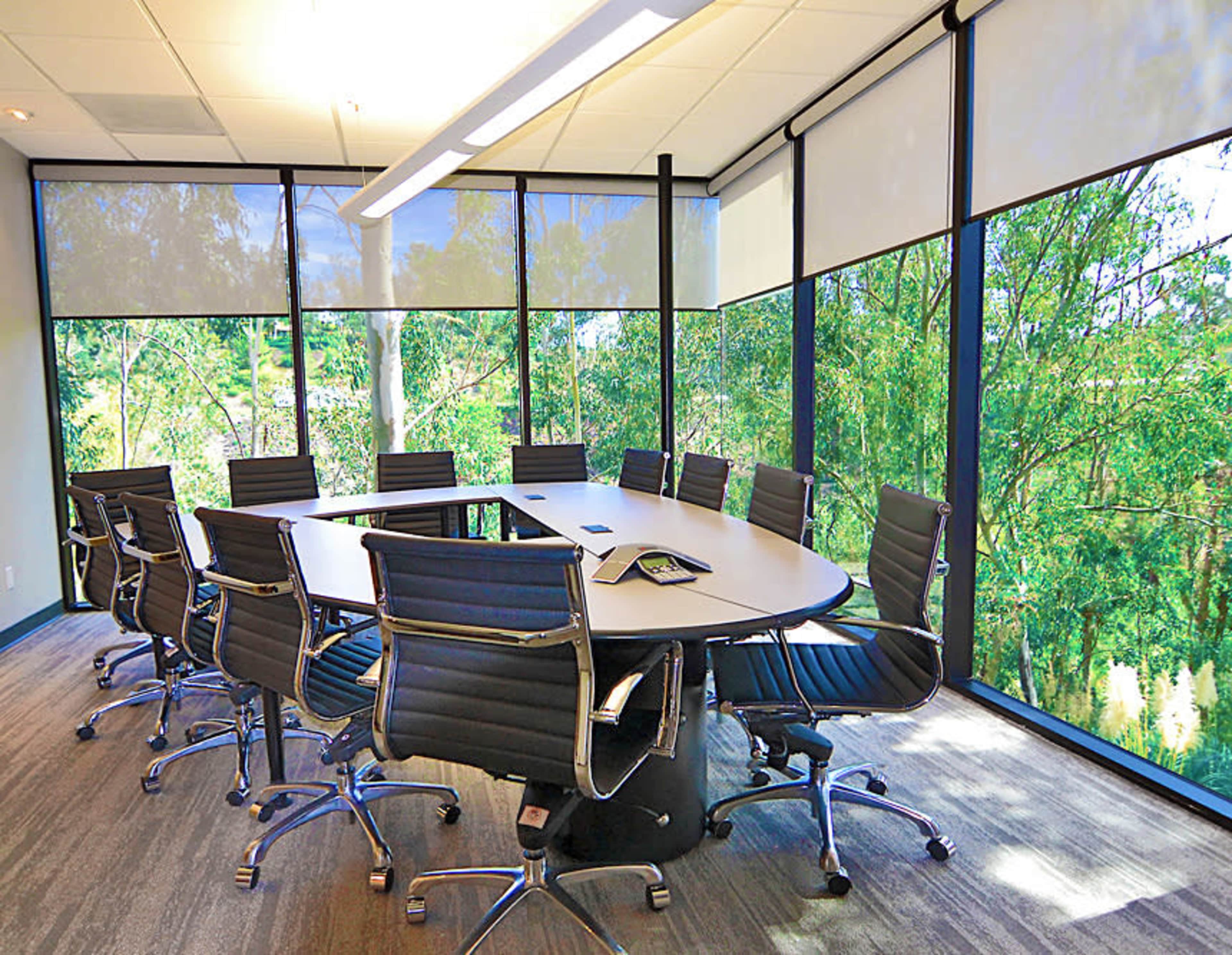 A conference room features a large oval table surrounded by black chairs and floor-to-ceiling windows showcasing greenery outside.