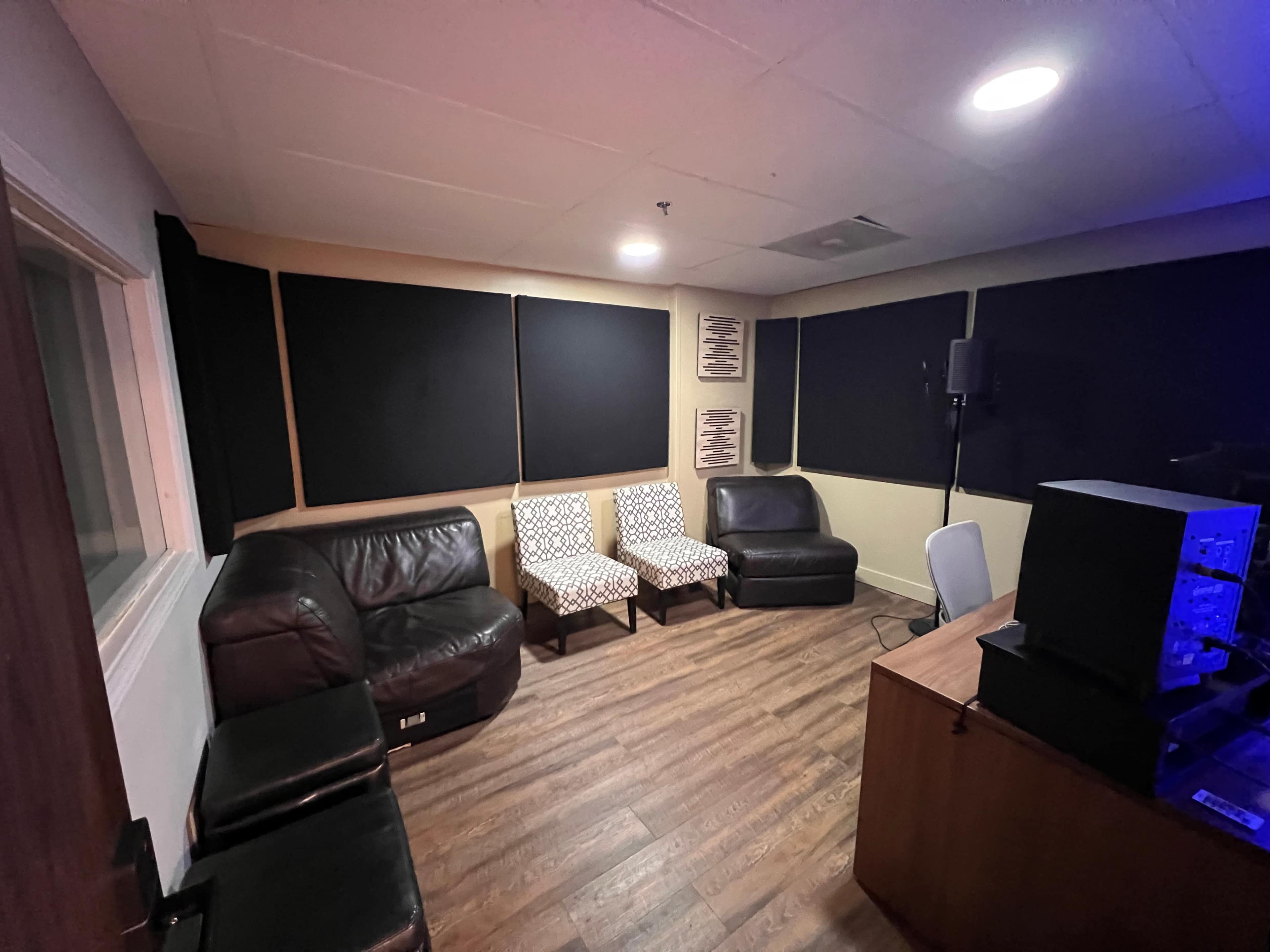 The image shows a small studio room with two patterned chairs, two leather couches, and soundproofing panels on the walls.