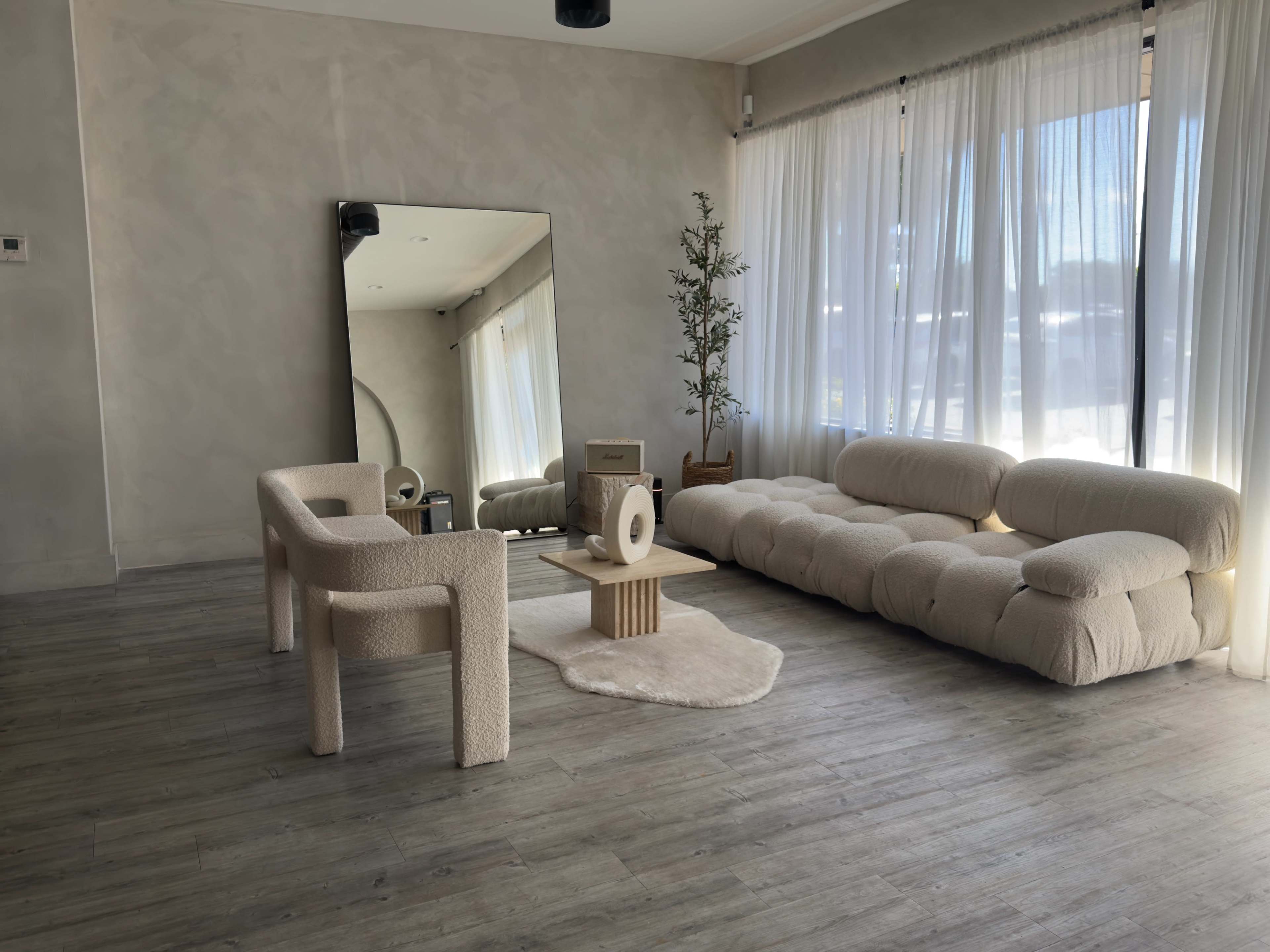 The image shows a modern living room with a light-colored sofa, a textured armchair, a round coffee table, and large windows letting in natural light.