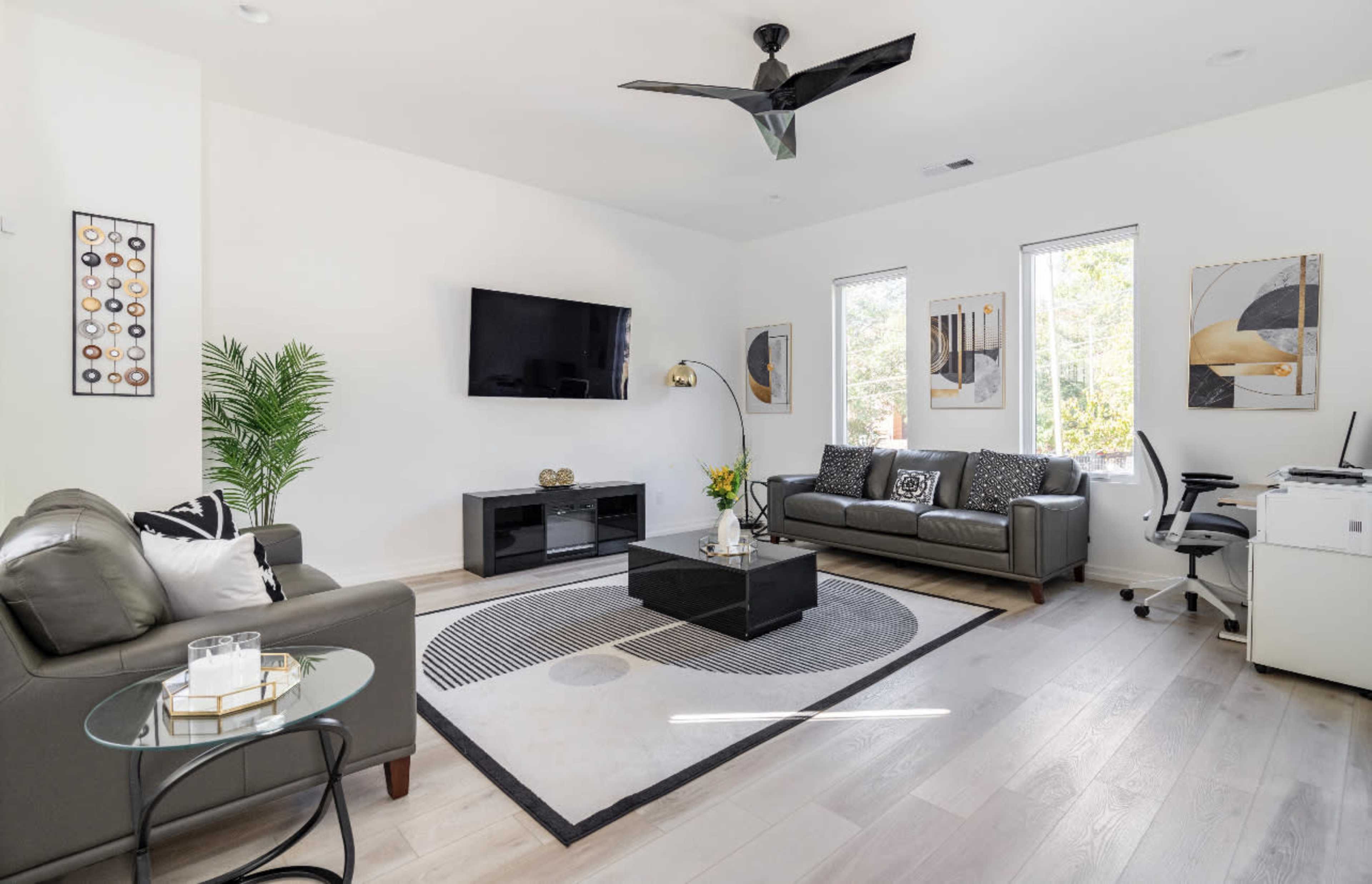 The living room features a large area rug, a black coffee table, a wall-mounted television, and modern furniture arranged around a window with natural light.