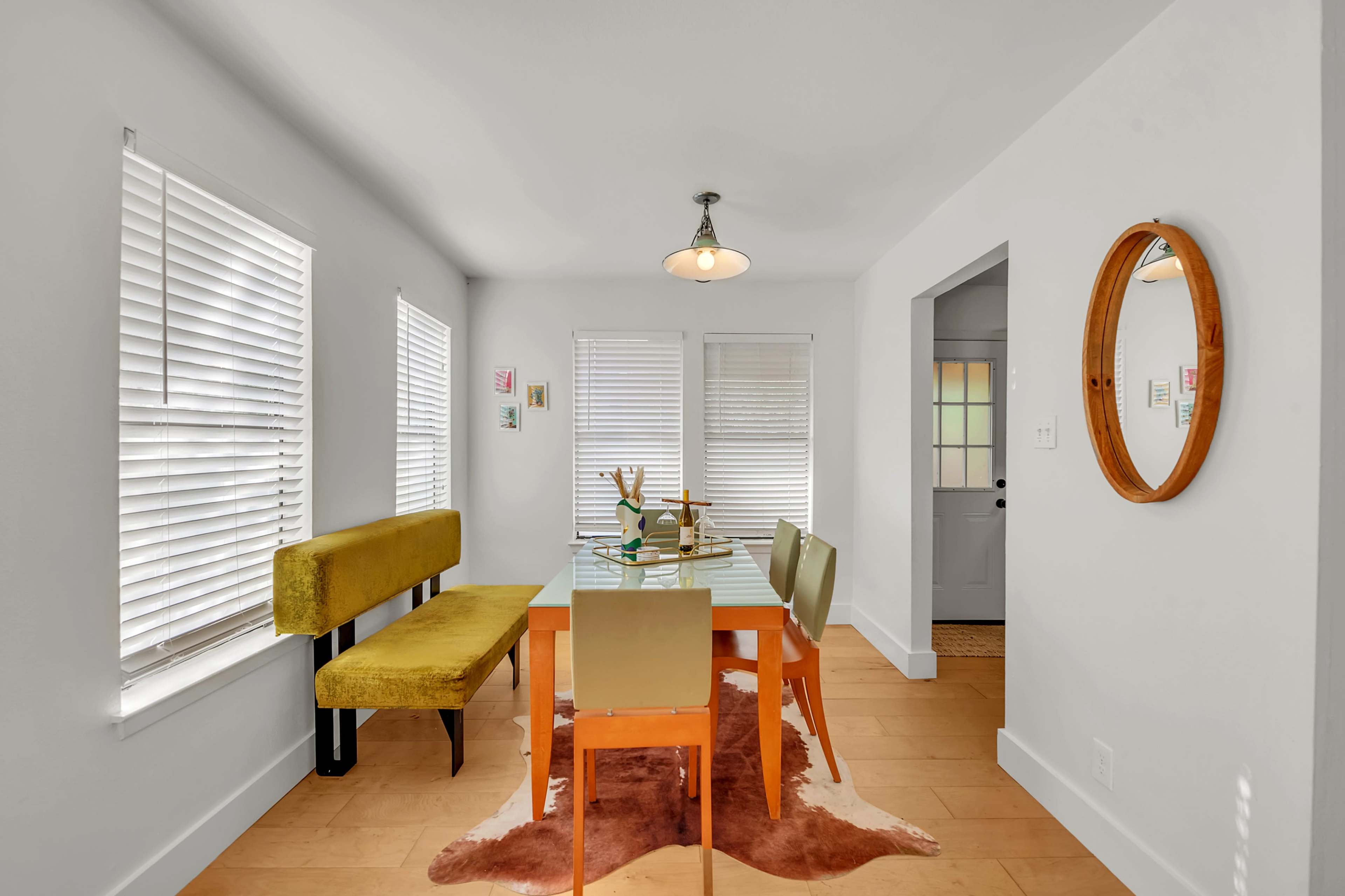 A dining area features a glass table surrounded by four chairs and a yellow bench, with windows partially covered by white blinds.