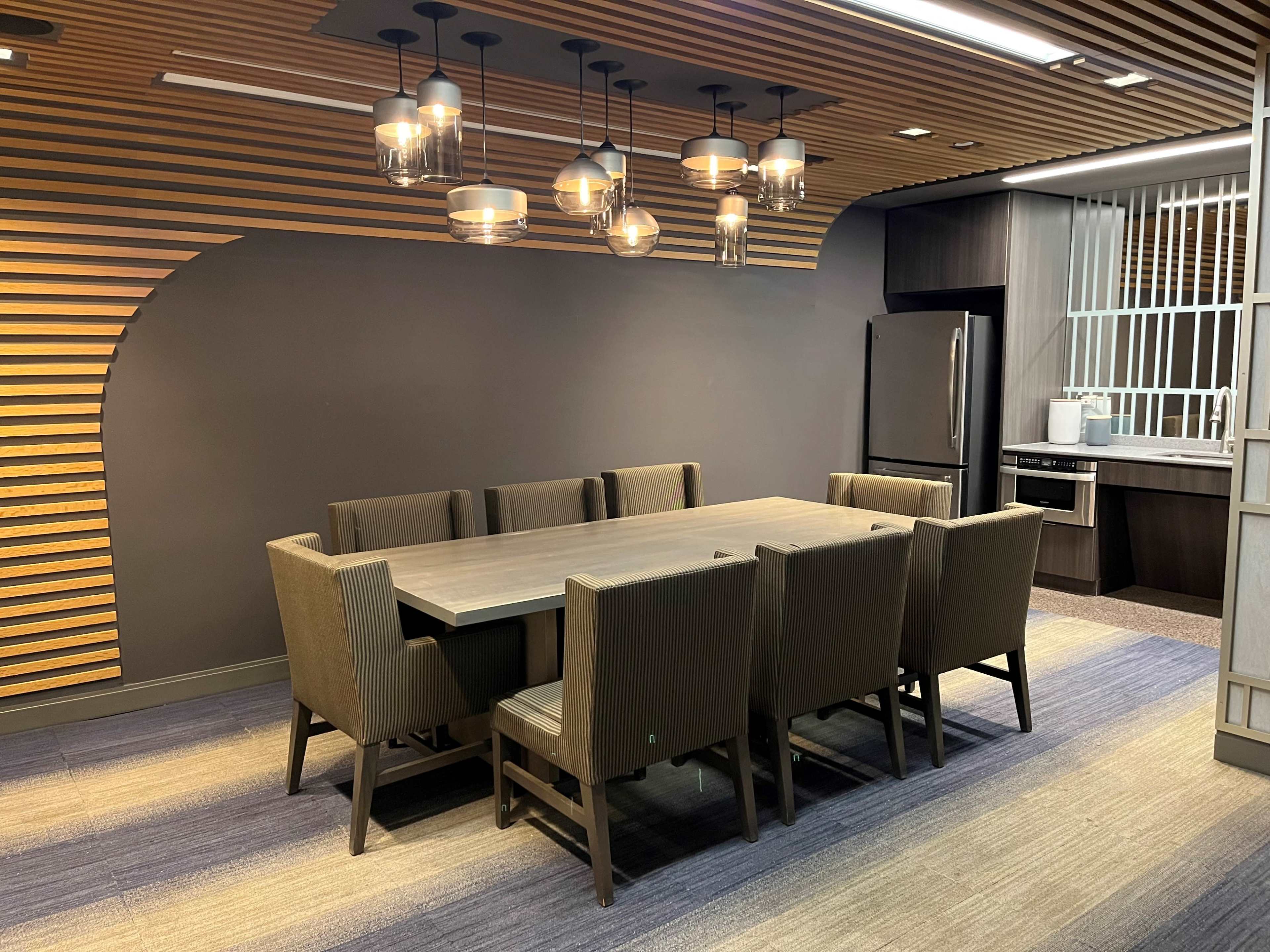 The image shows a modern conference room with a long wooden table surrounded by eight upholstered chairs, illuminated by pendant lights, and featuring a dark accent wall and a kitchenette in the background.