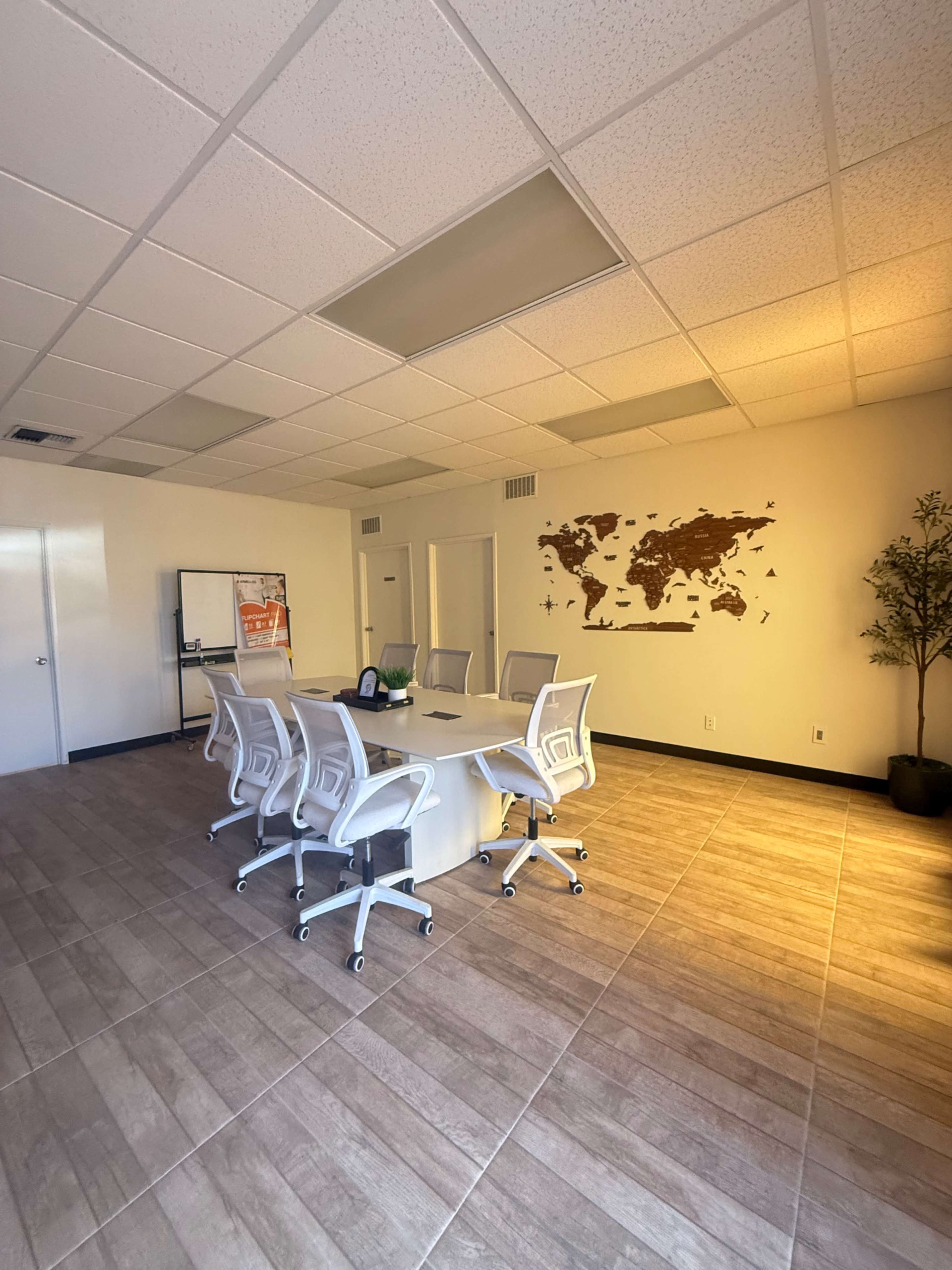 A conference room features a round table surrounded by white chairs, with a world map on the wall and a large window allowing natural light.