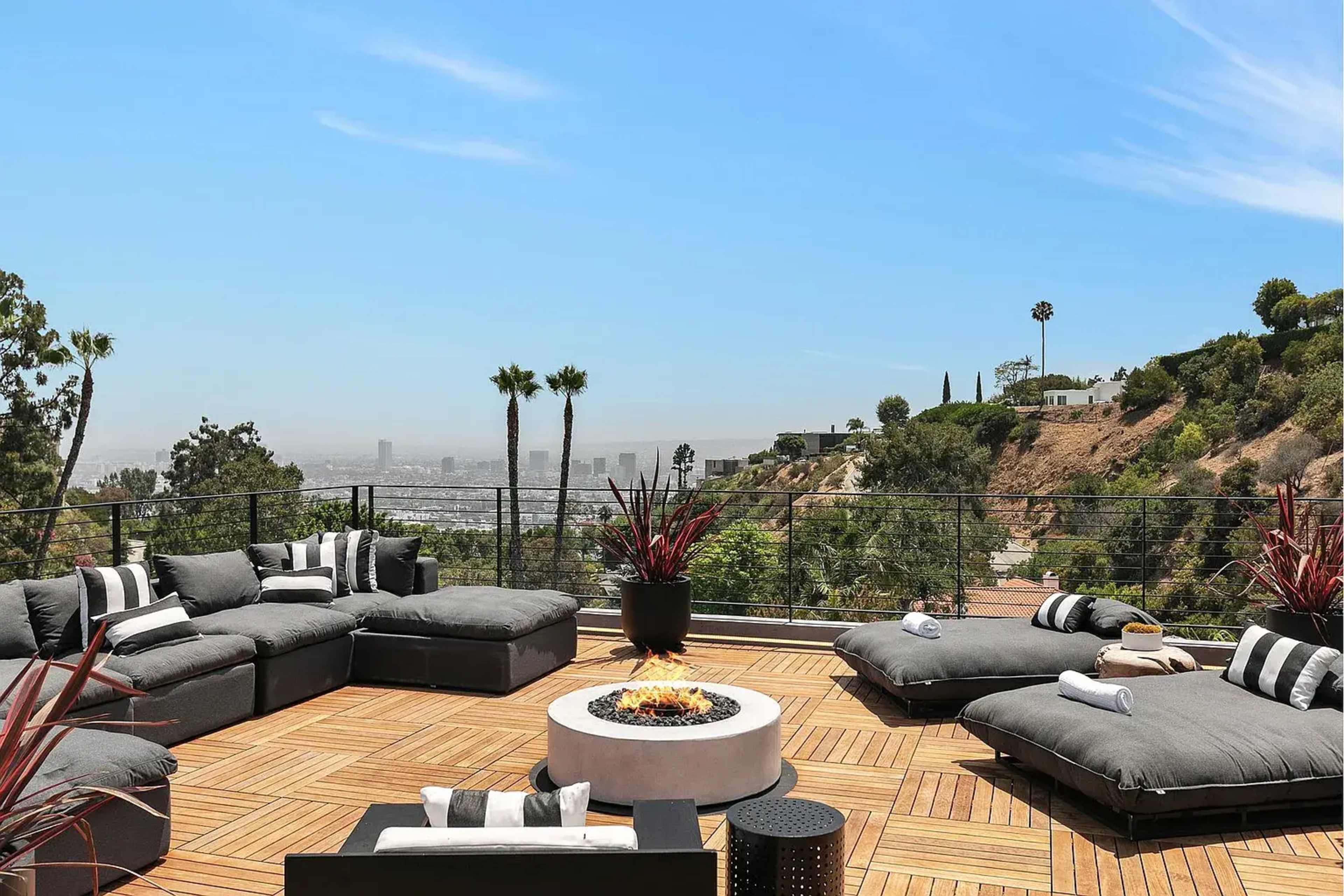 A stylish outdoor patio features gray seating, a fire pit, and views of the city skyline surrounded by palm trees and hills.