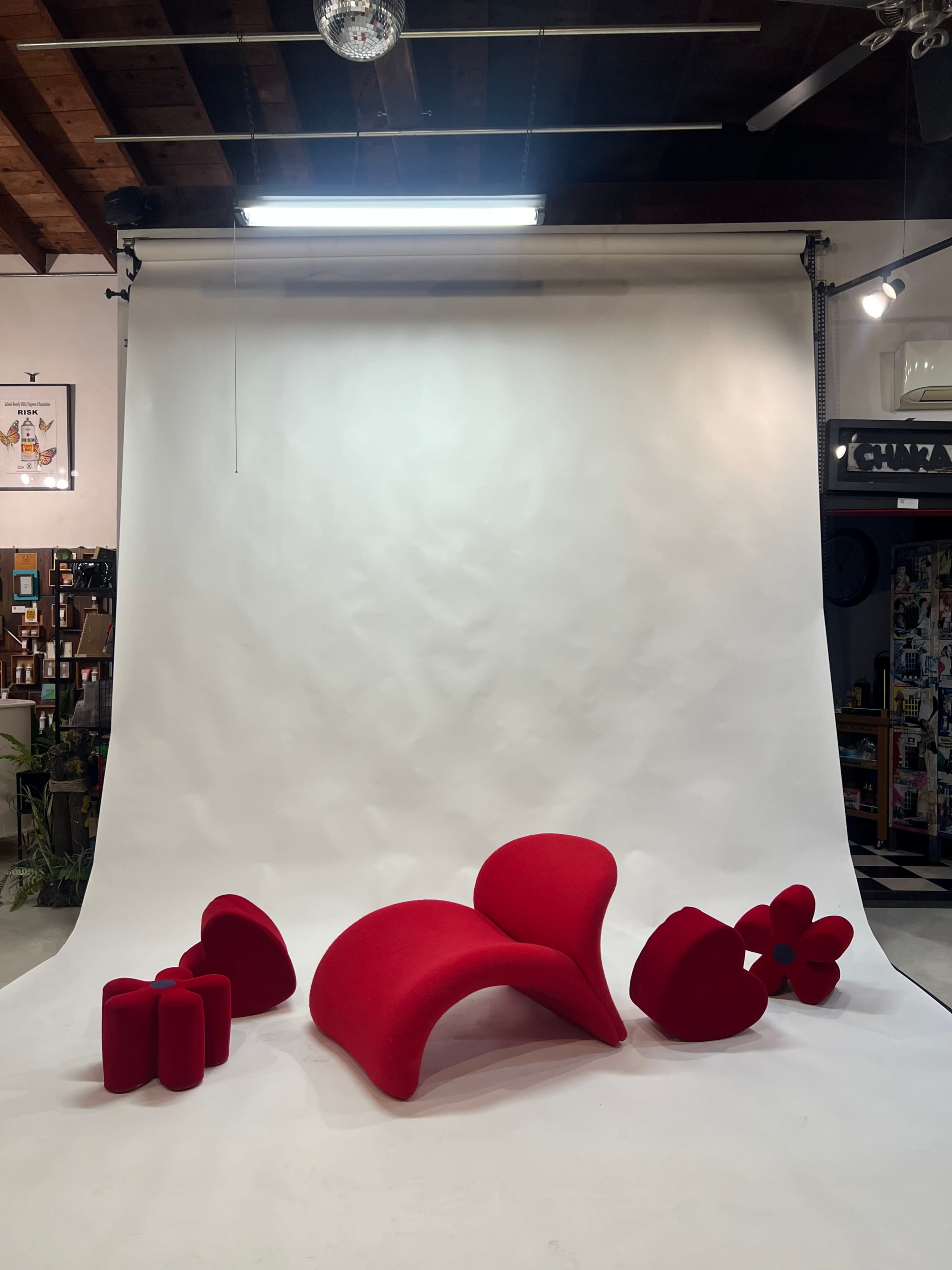 The image features a minimalist studio setting with a white backdrop and several abstract red furniture pieces arranged on the floor.