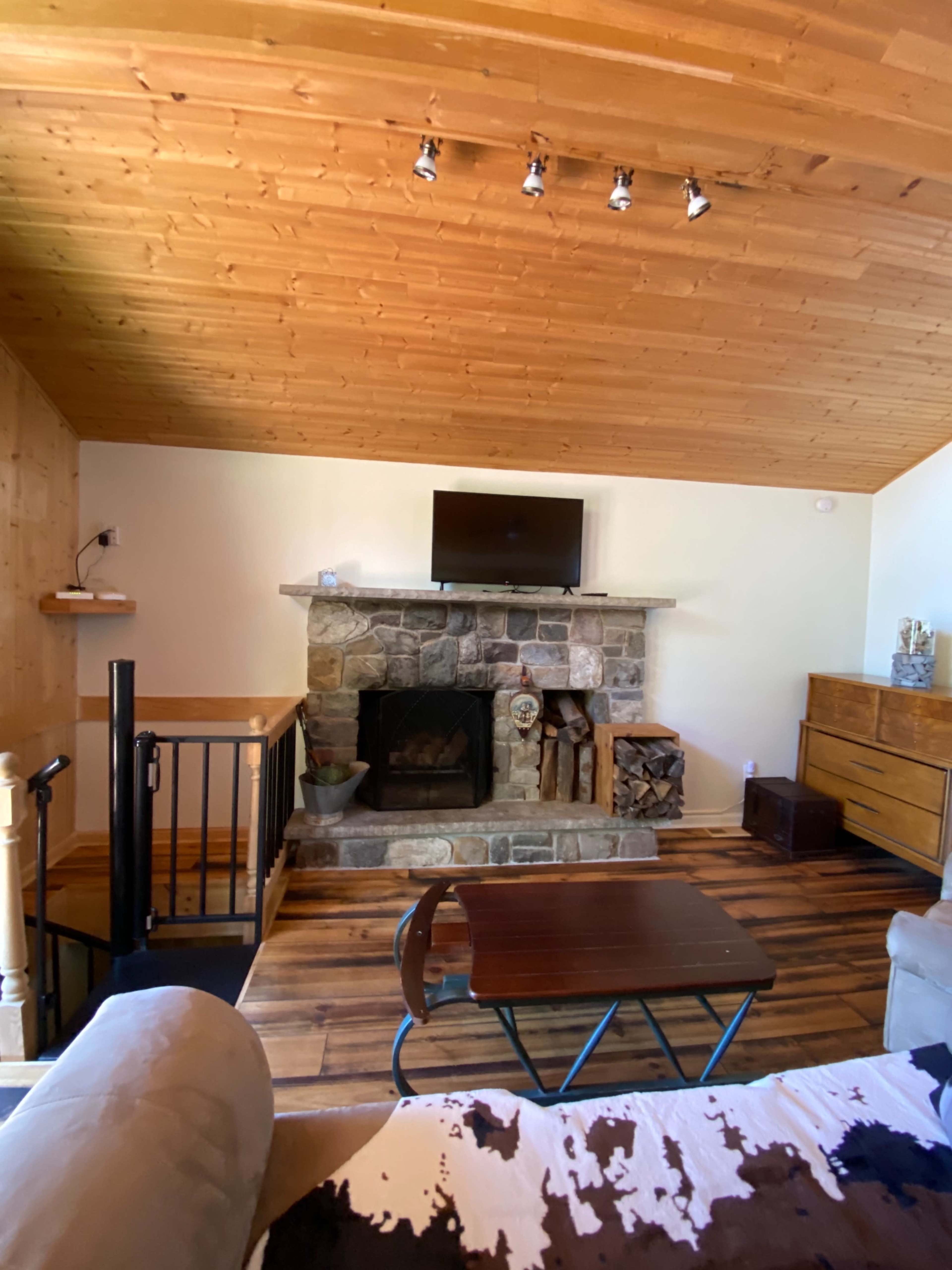 The image shows a living room with a stone fireplace, a flat-screen TV mounted above it, and wooden flooring, along with a staircase and a wooden dresser.