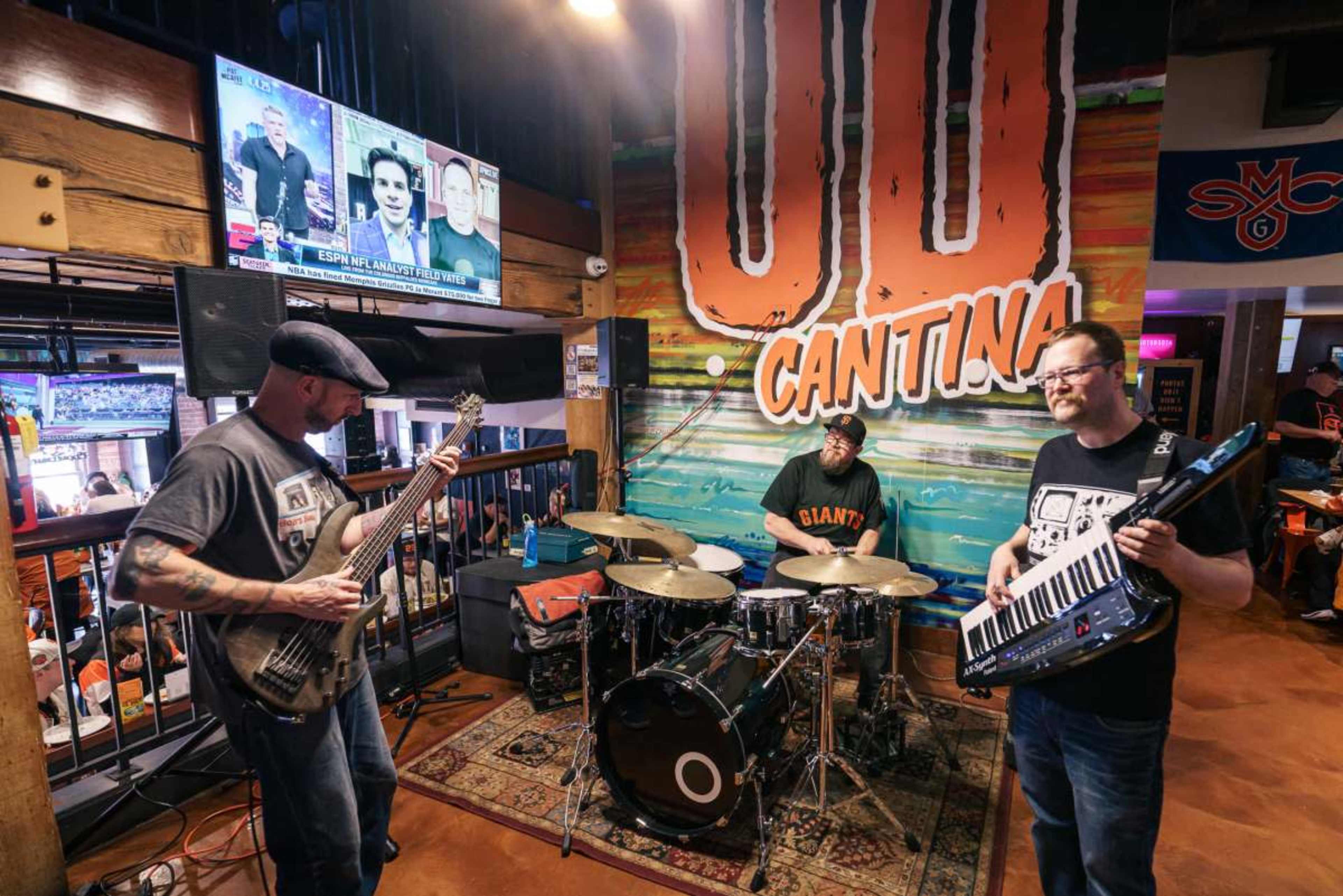 A live band performs in a lively cantina, with a guitarist, drummer, and keyboard player as patrons watch a sports event on a nearby television.