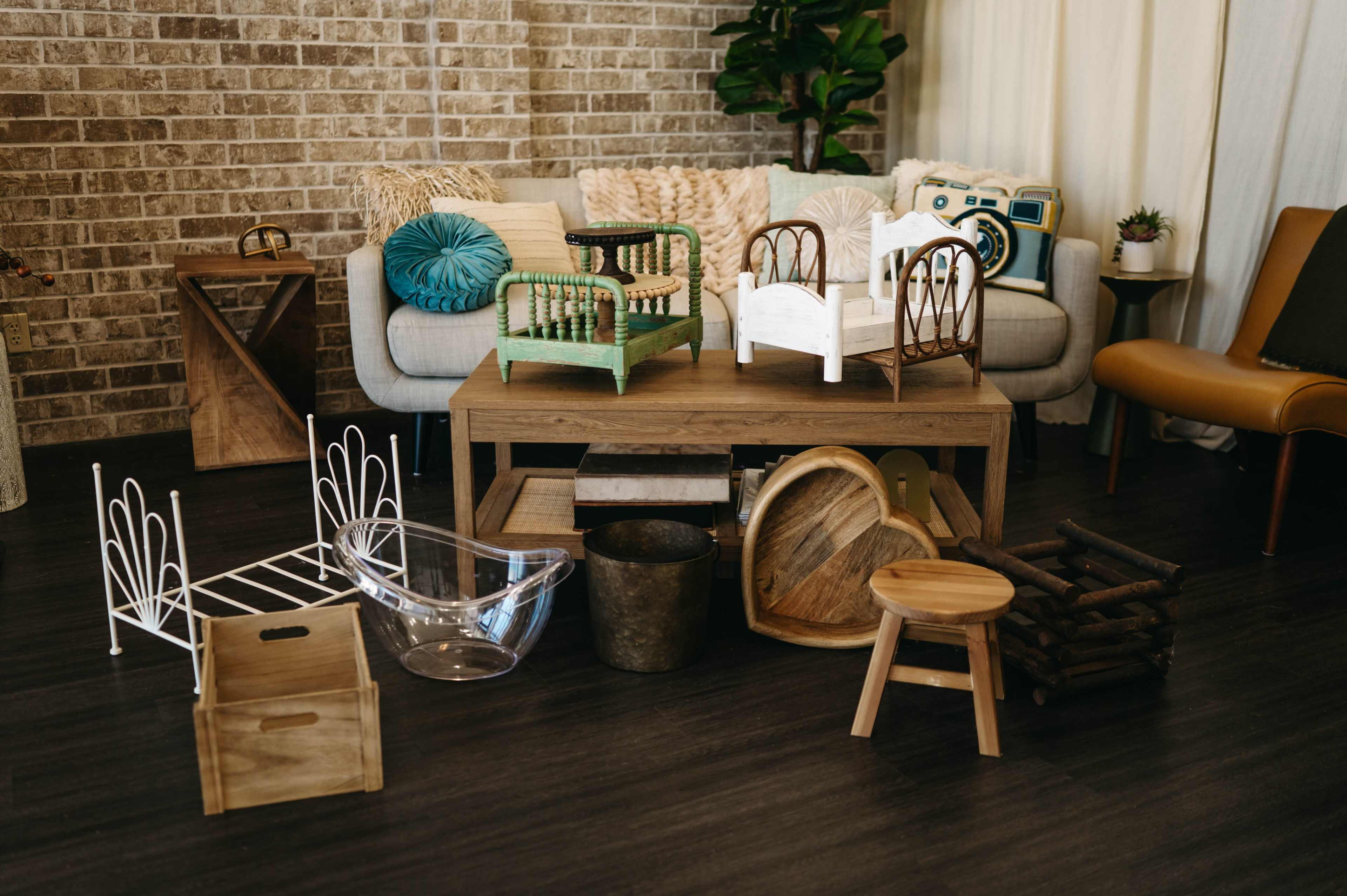 The image features a variety of furniture and decorative items arranged on a dark floor, including small beds, a wooden table, a glass container, and a potted plant, against a brick wall backdrop.
