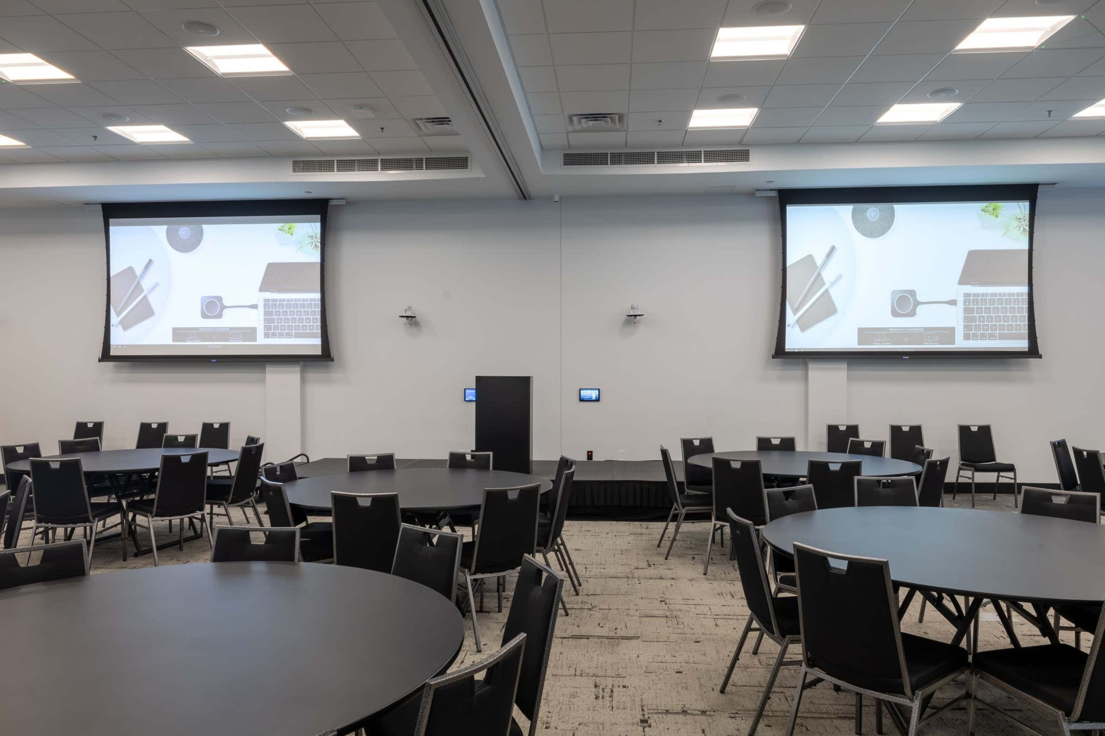 A conference room features multiple round tables and two large projection screens displaying computer-related graphics.