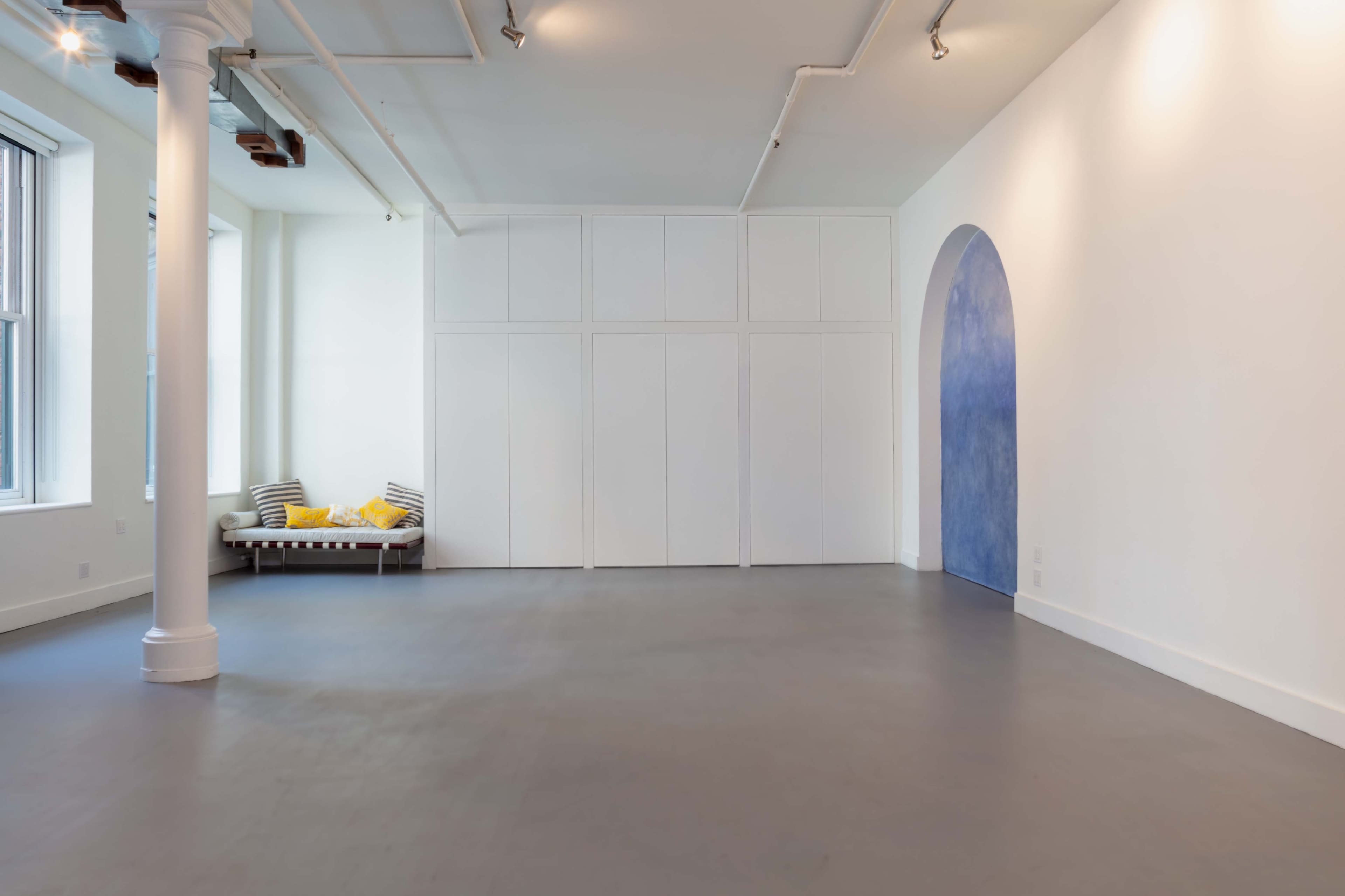 A spacious, minimalist room with a light gray floor, white walls, and a blue archway, featuring a seating area with yellow cushions along one side.