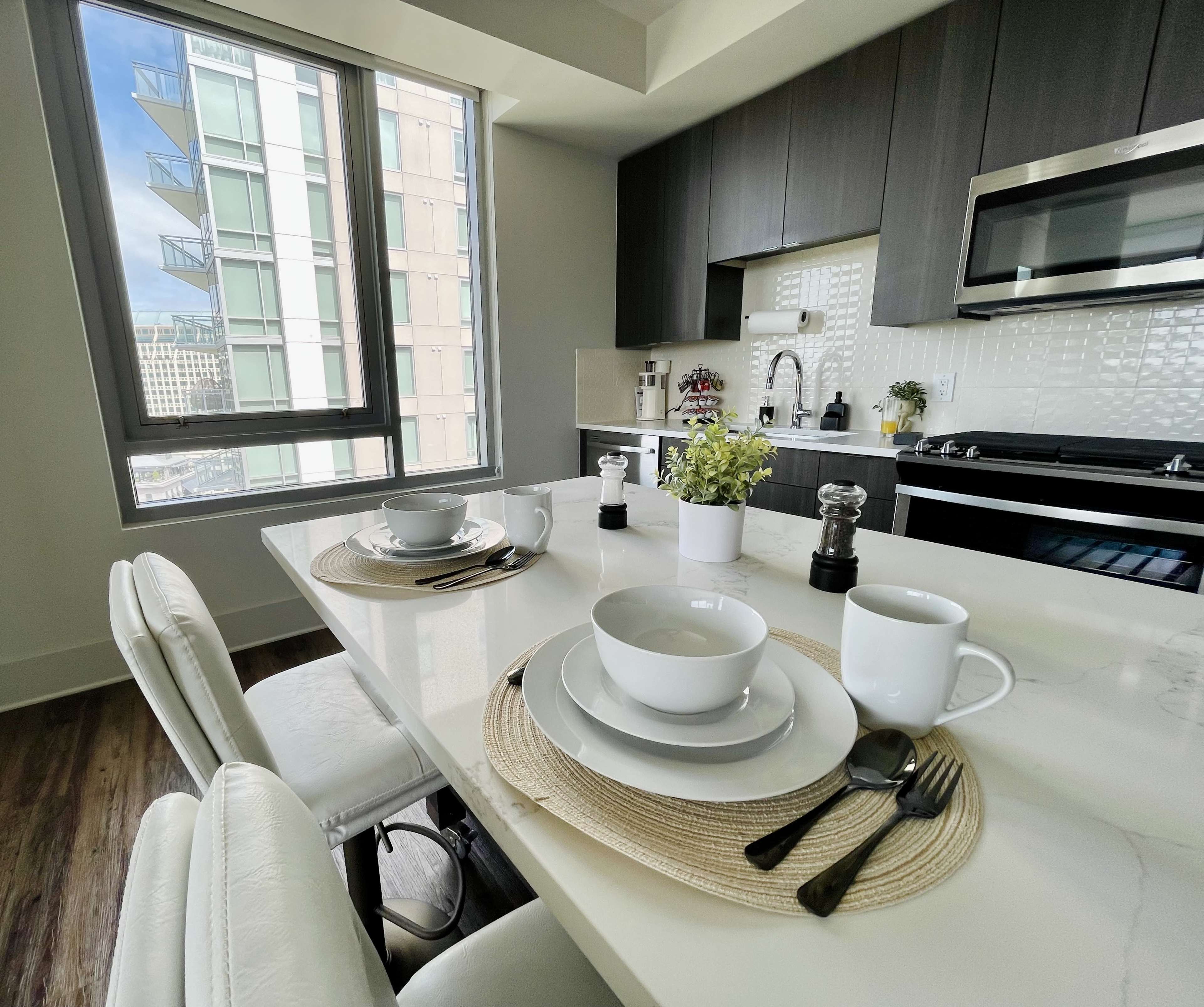 A modern kitchen features a dining table set with plates, cups, and utensils, alongside a view of the city through a large window.