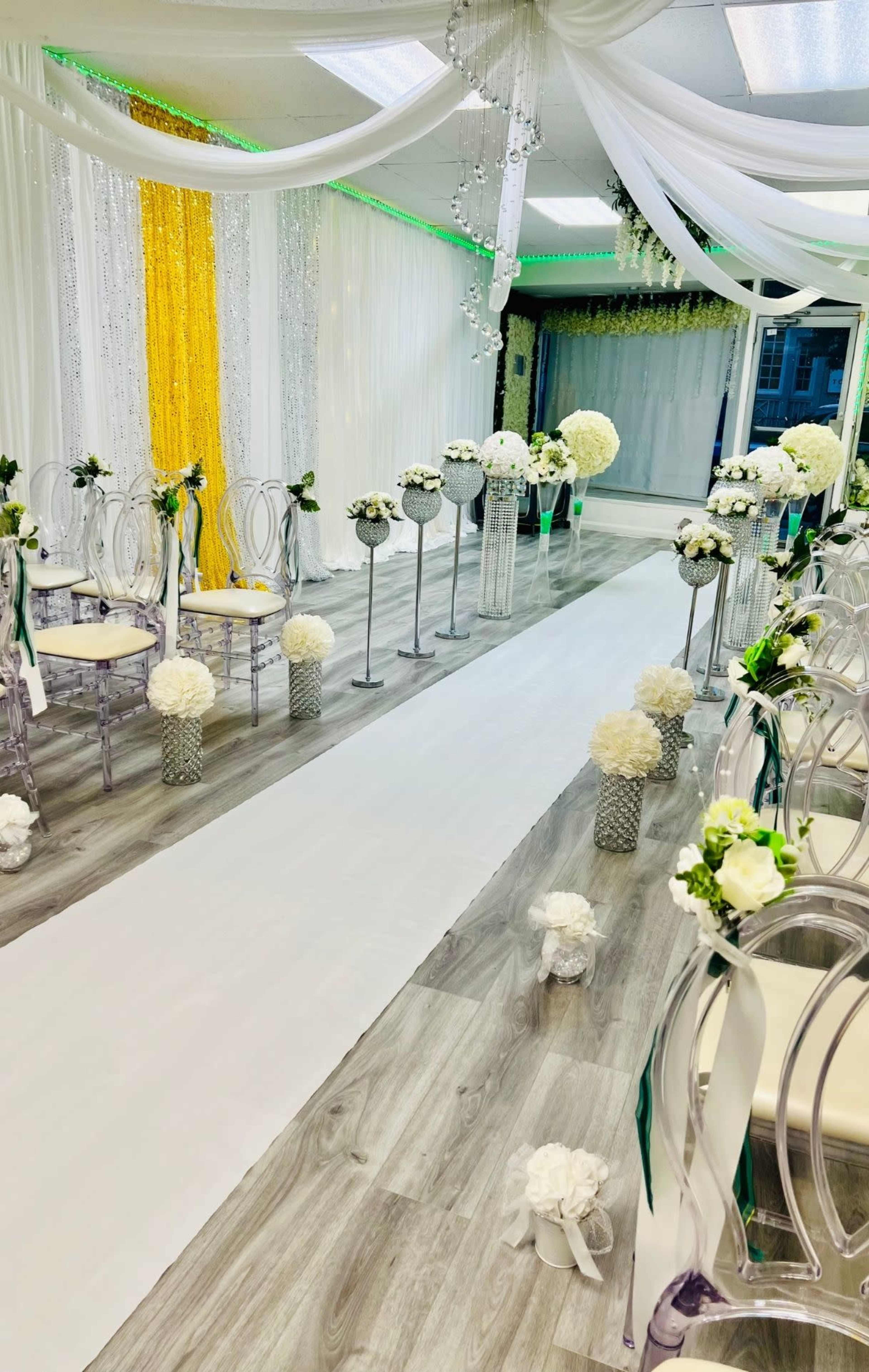 The image shows a decorated indoor venue with an aisle lined with white runners, floral arrangements, and elegant seating ready for a ceremony.