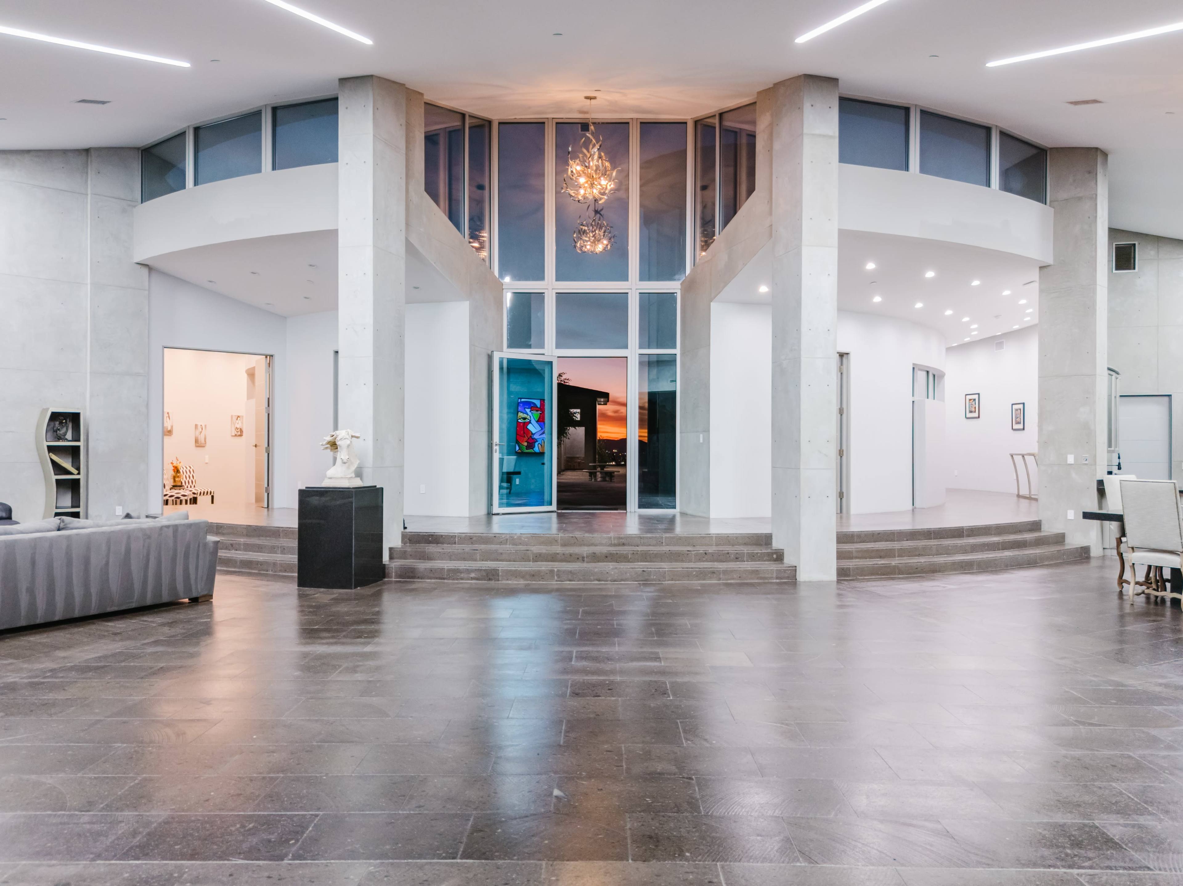 The image shows a spacious, modern interior with high ceilings, large windows, and a staircase leading to a central entryway.