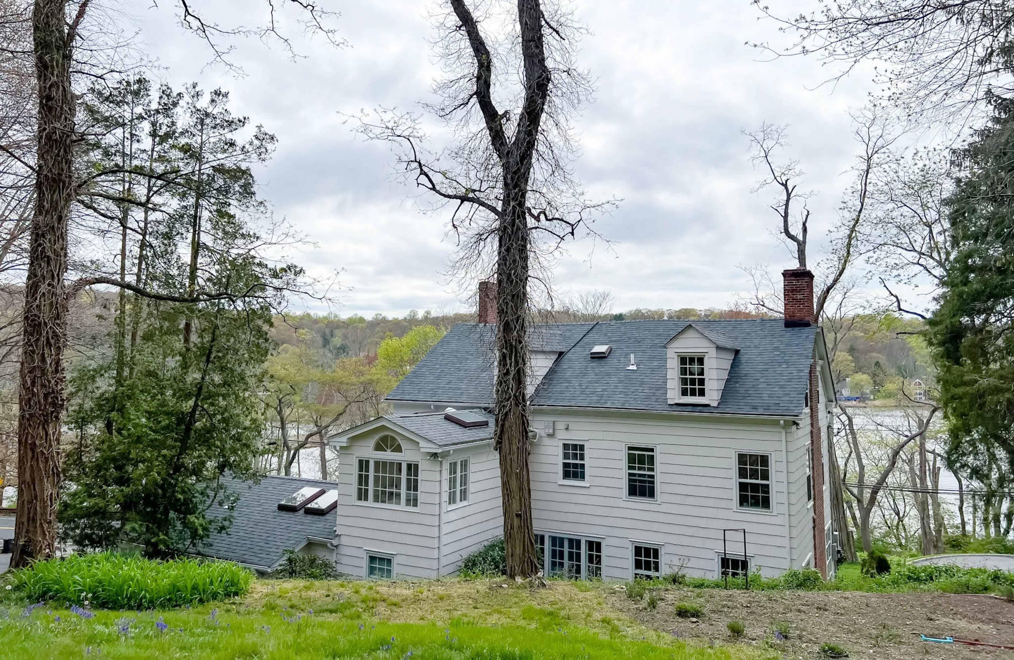 A white two-story house is situated on a hillside overlooking a body of water, surrounded by trees and greenery.