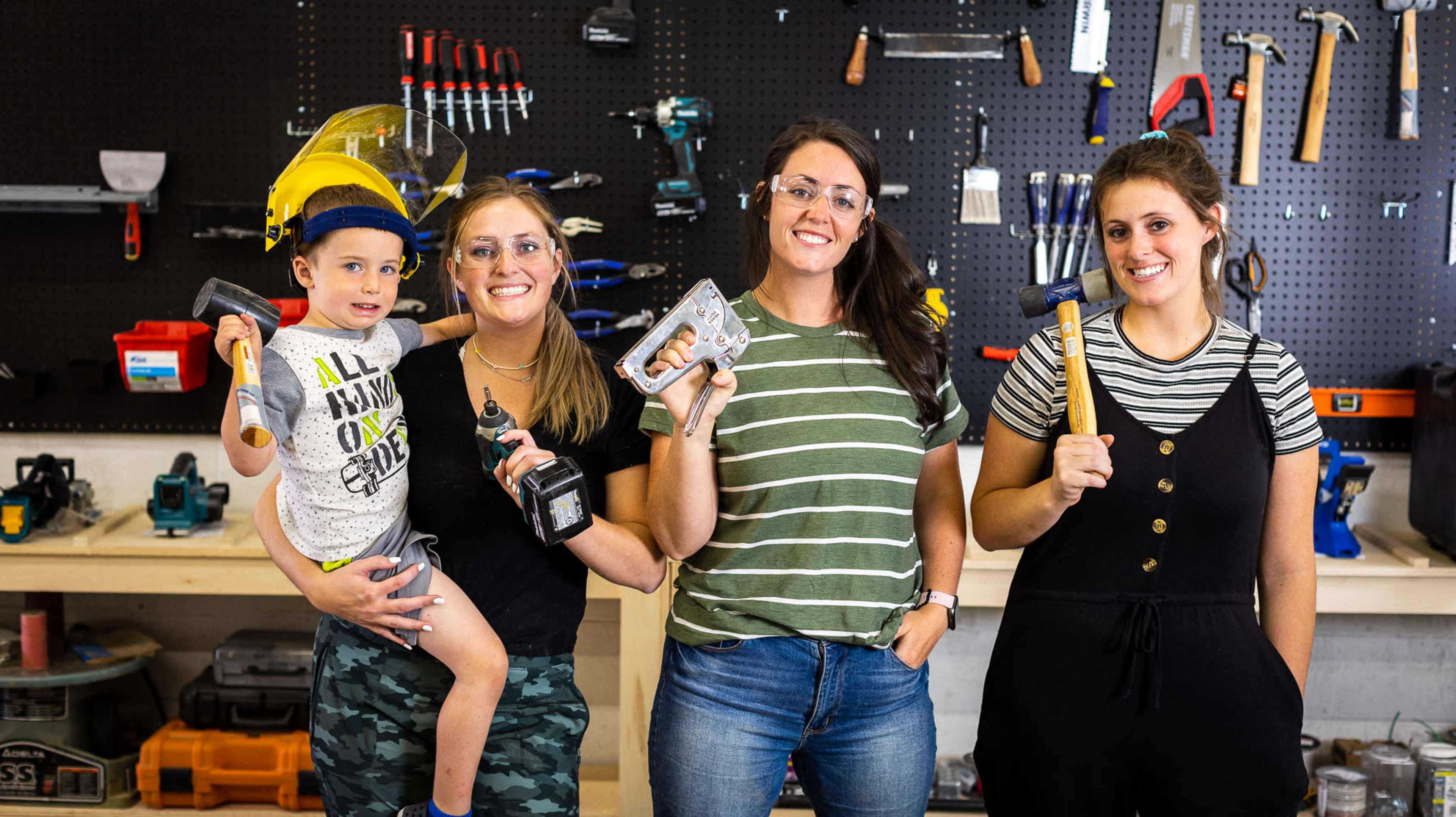 Four people stand in a workshop, with one child being held, as they each hold tools and wear safety glasses.