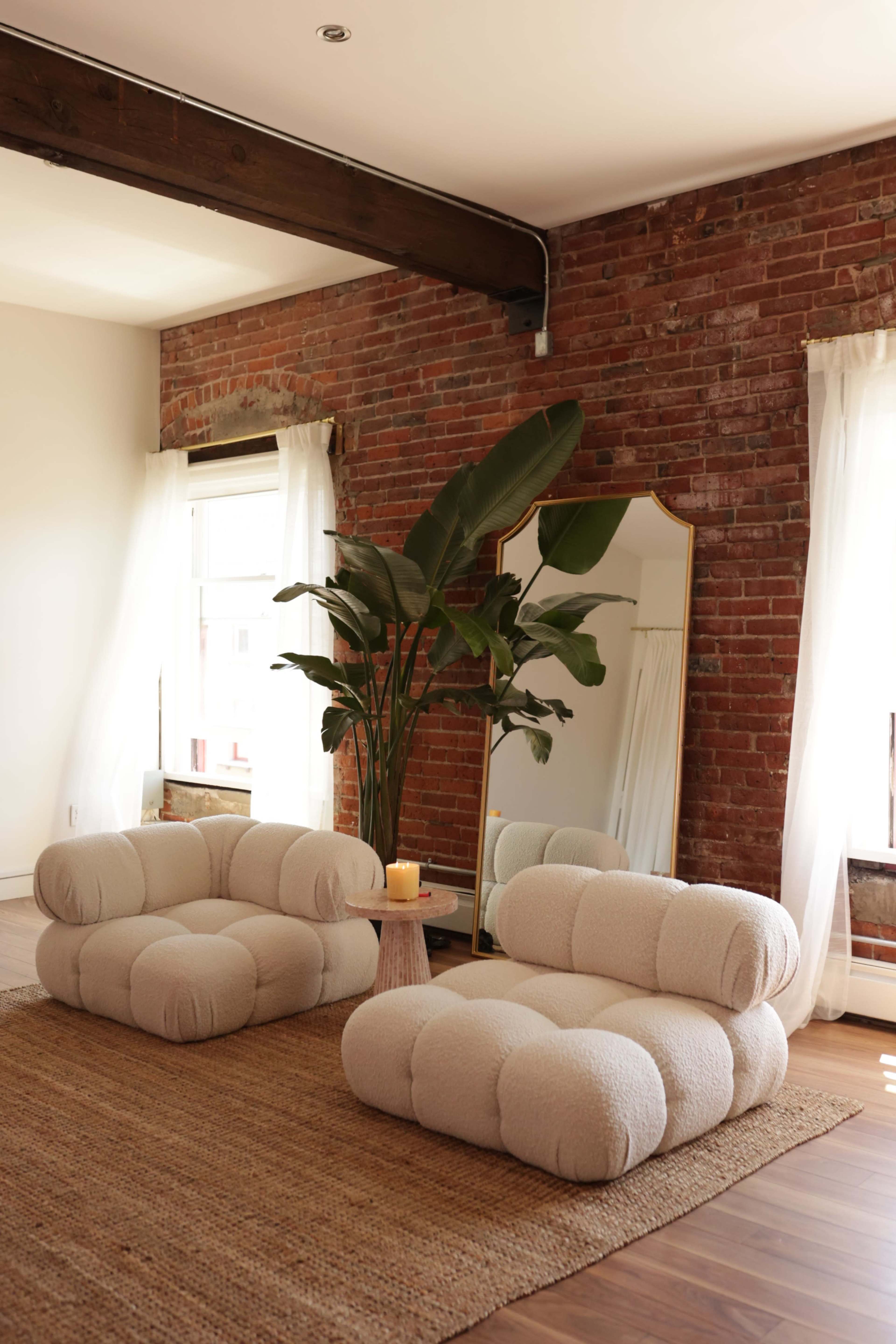 The image features a cozy living area with two plush, cream-colored chairs, a large mirror, a potted plant, and natural light streaming through white curtains.