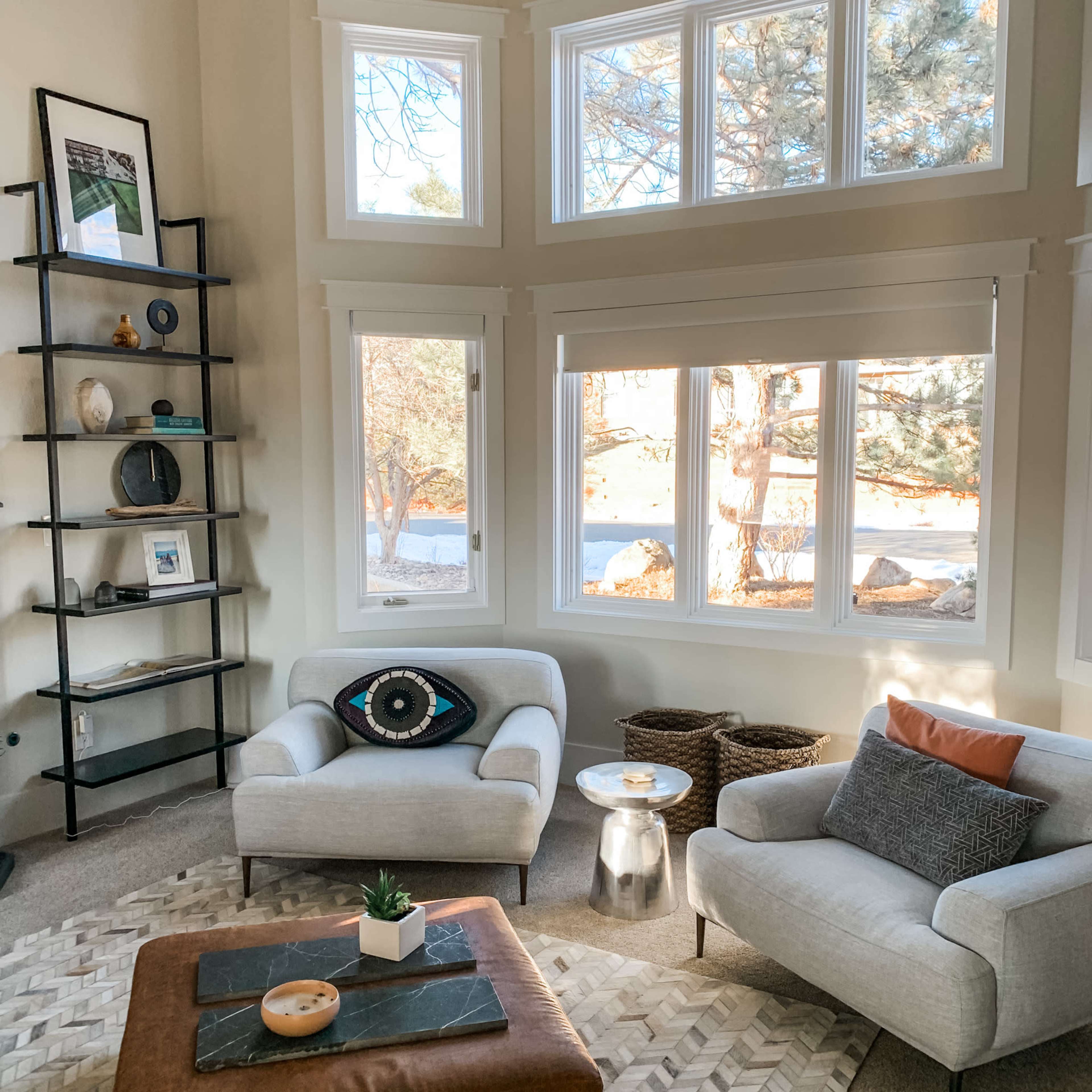 The image depicts a cozy living room with large windows, two stylish couches, a small side table, and a shelf displaying decorative items.