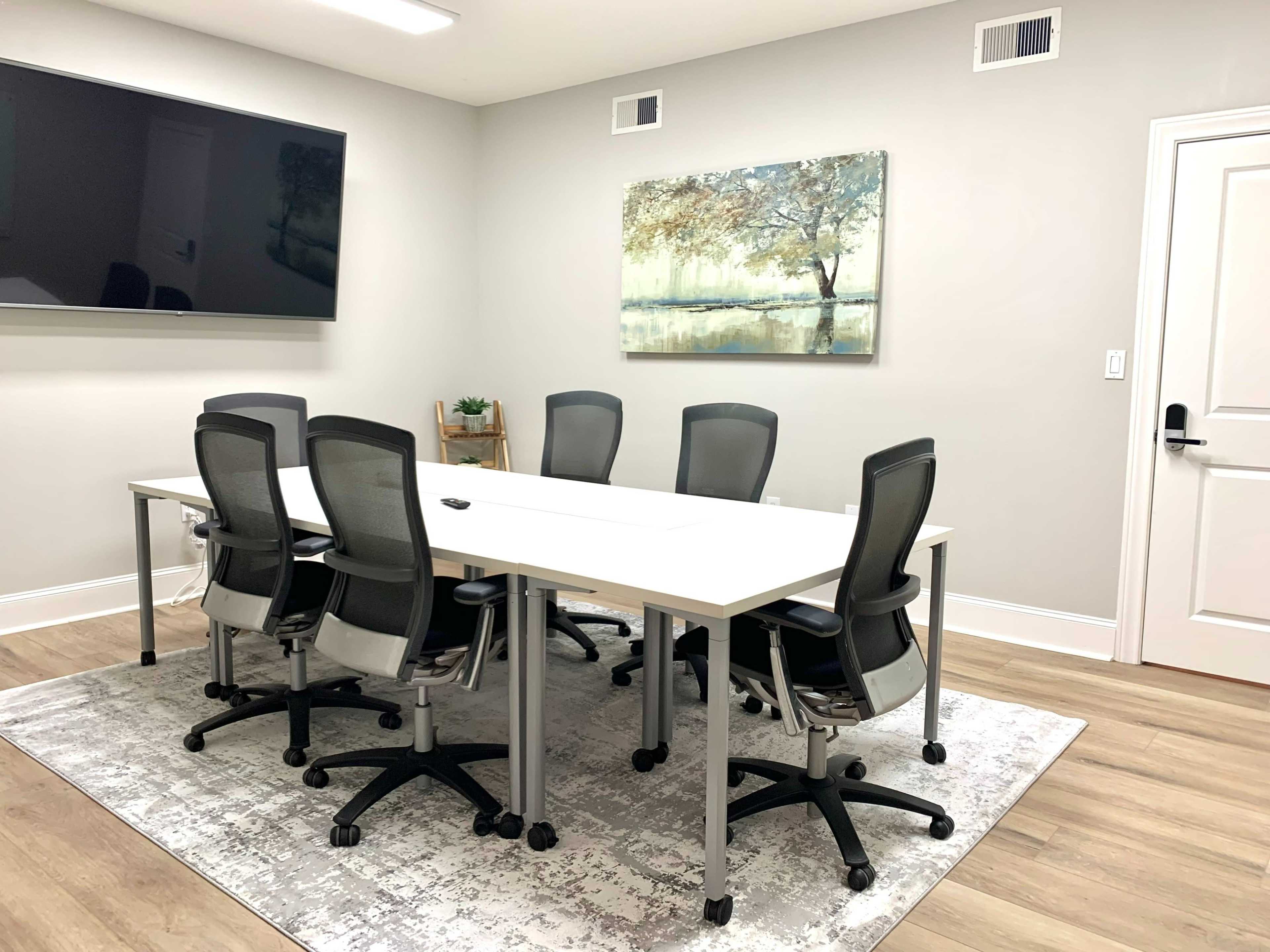 A conference room features a long white table surrounded by six gray office chairs, a large screen on the wall, and an abstract landscape painting.