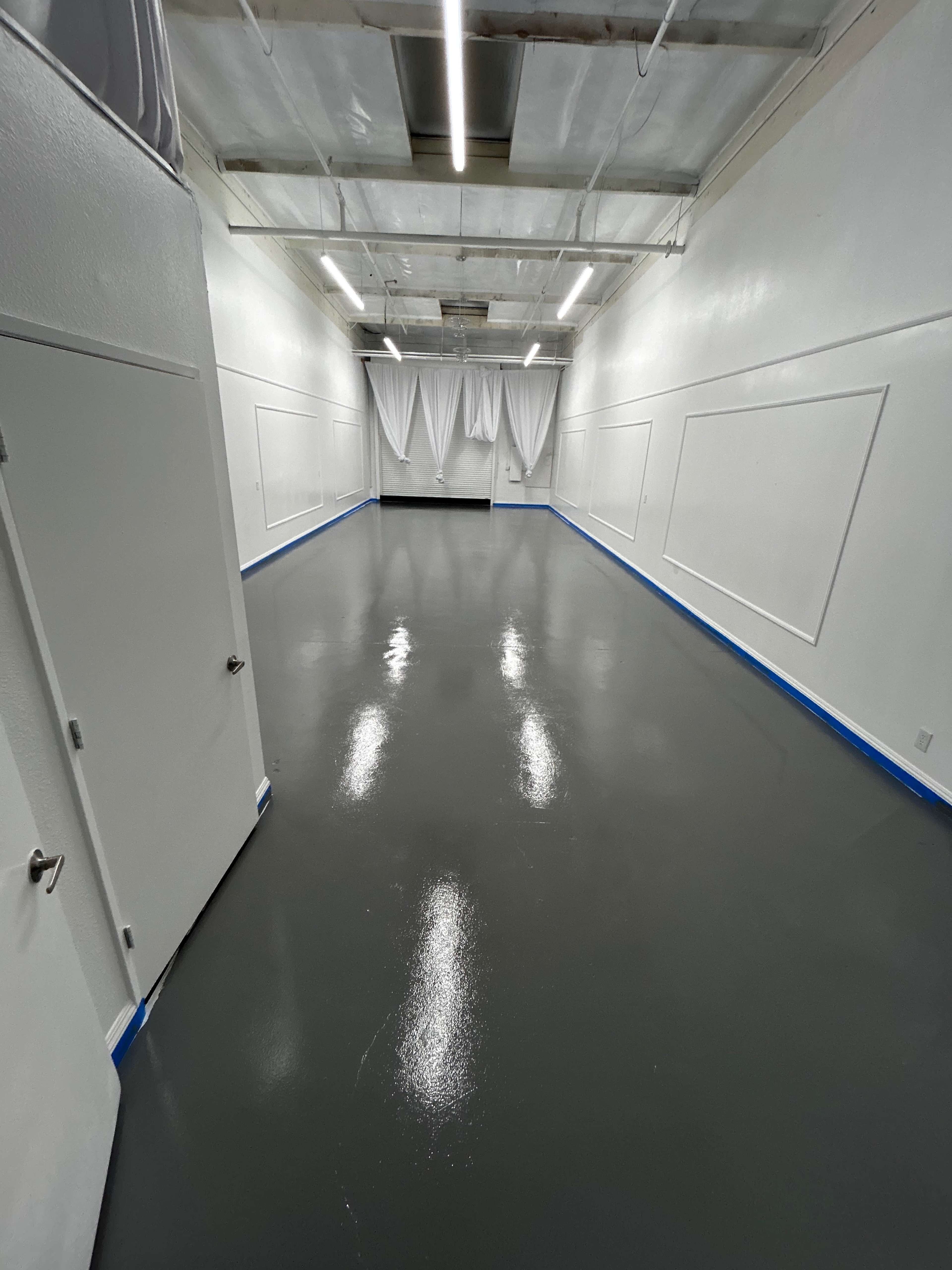 A spacious, empty room with a glossy dark gray floor, white walls, and partially closed curtains at one end.