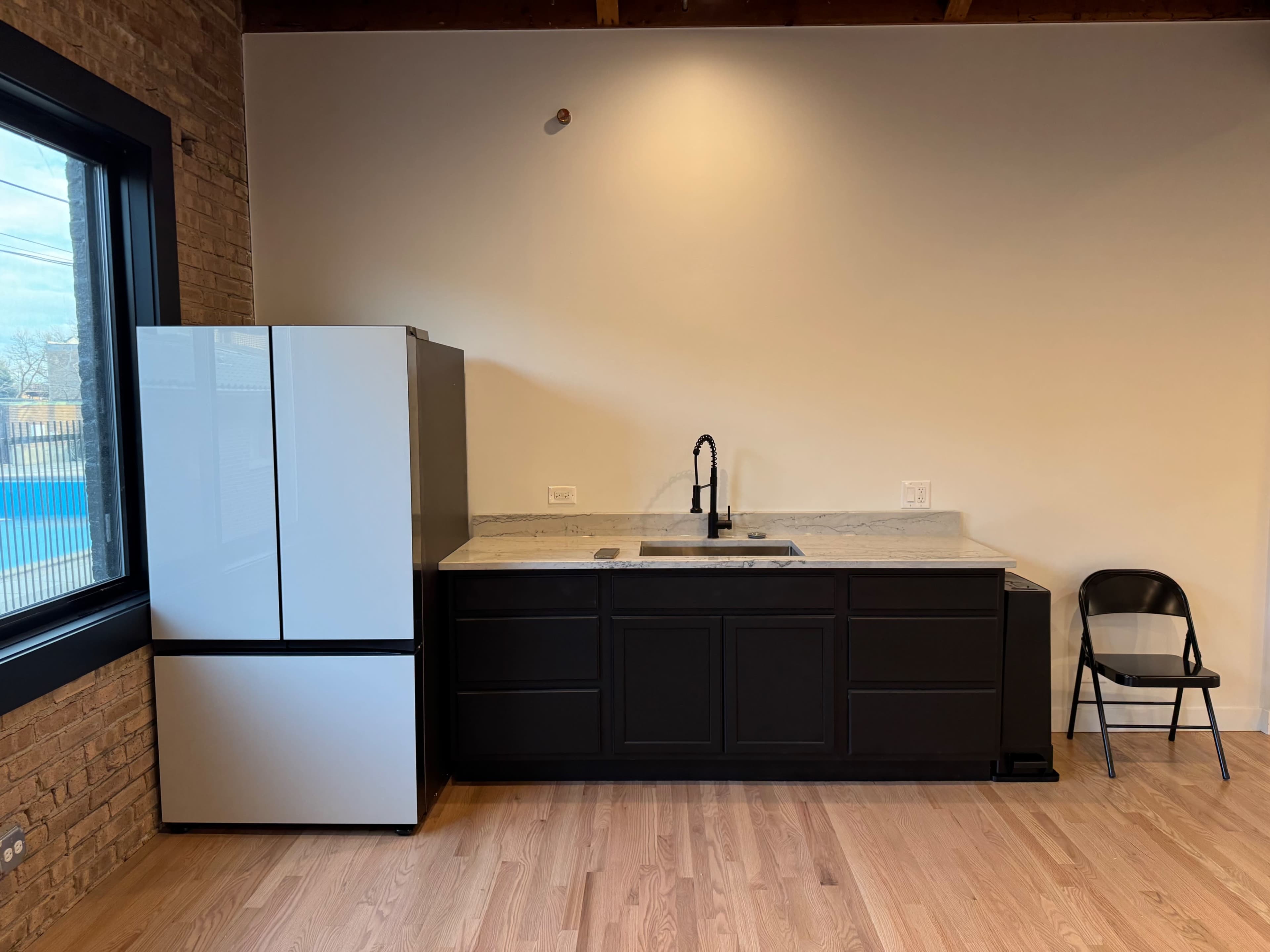 The image shows a modern kitchen featuring a black cabinetry, a white refrigerator, a sink with a faucet, and a single black chair against a wall with exposed brick.