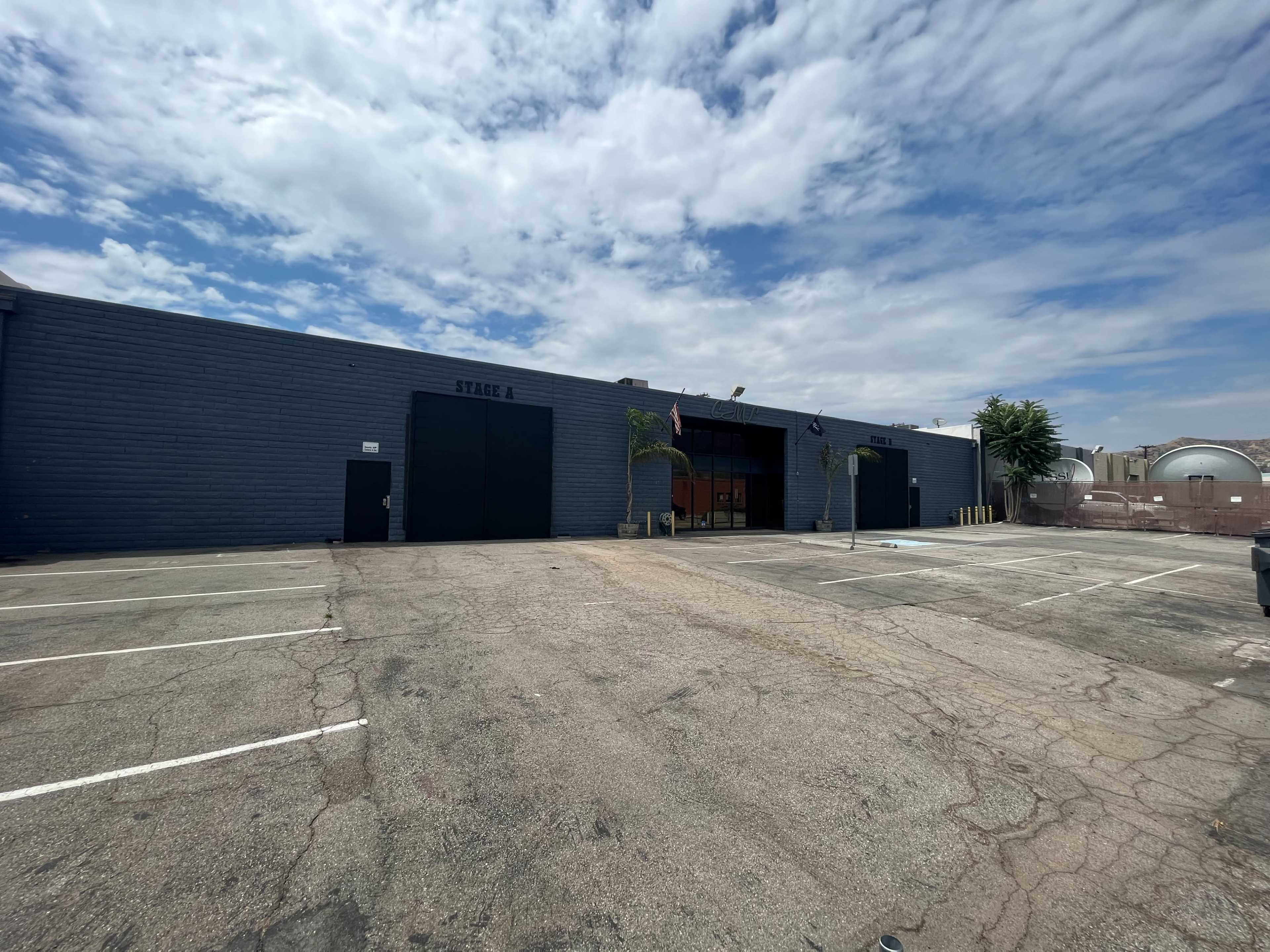 The image shows a large, gray building labeled "STAGE 8" with a parking lot in front and cloudy skies overhead.