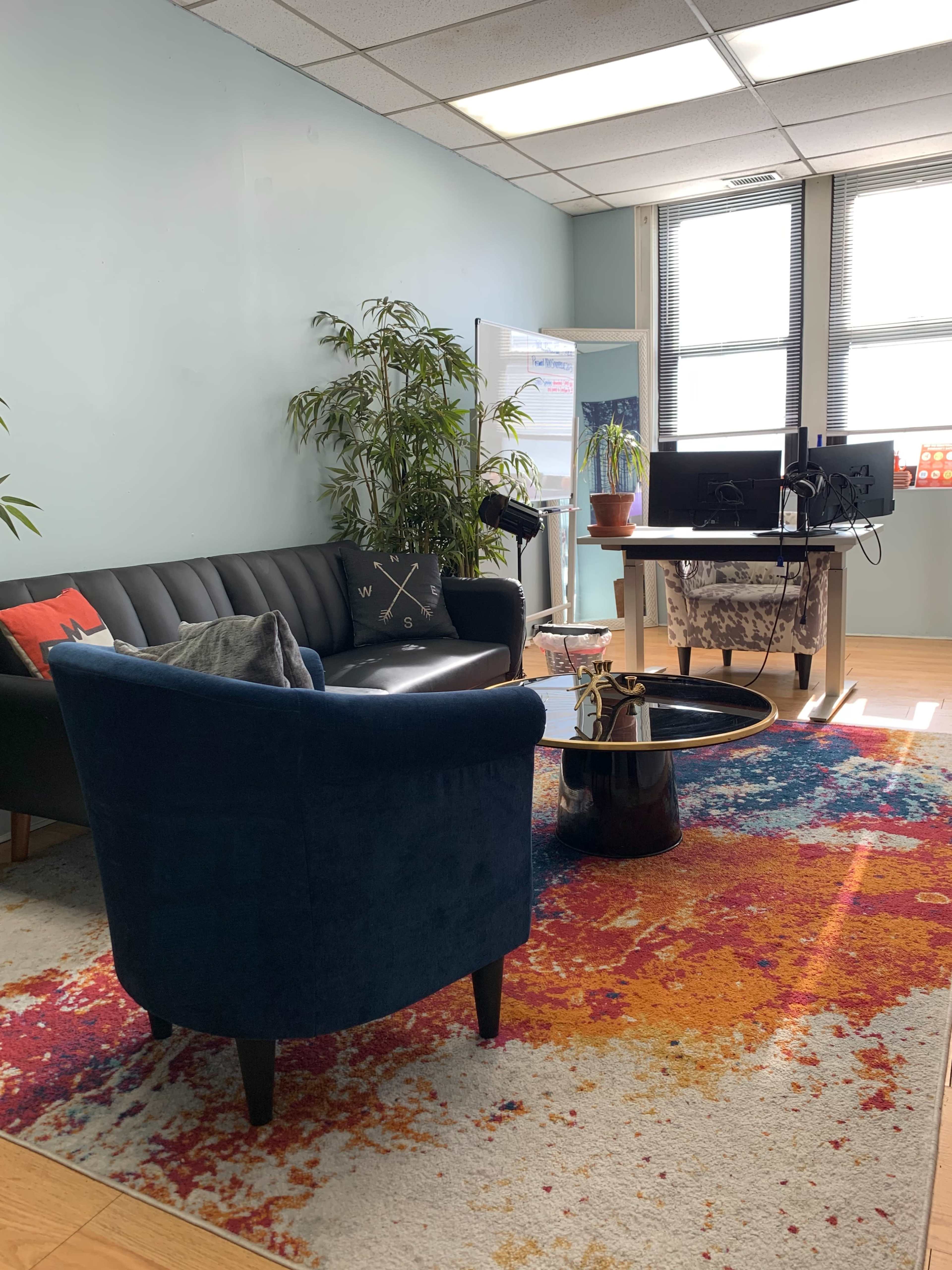 A modern office space with a blue couch, a chair, a round coffee table, and a desk with two monitors, all arranged on a colorful rug.