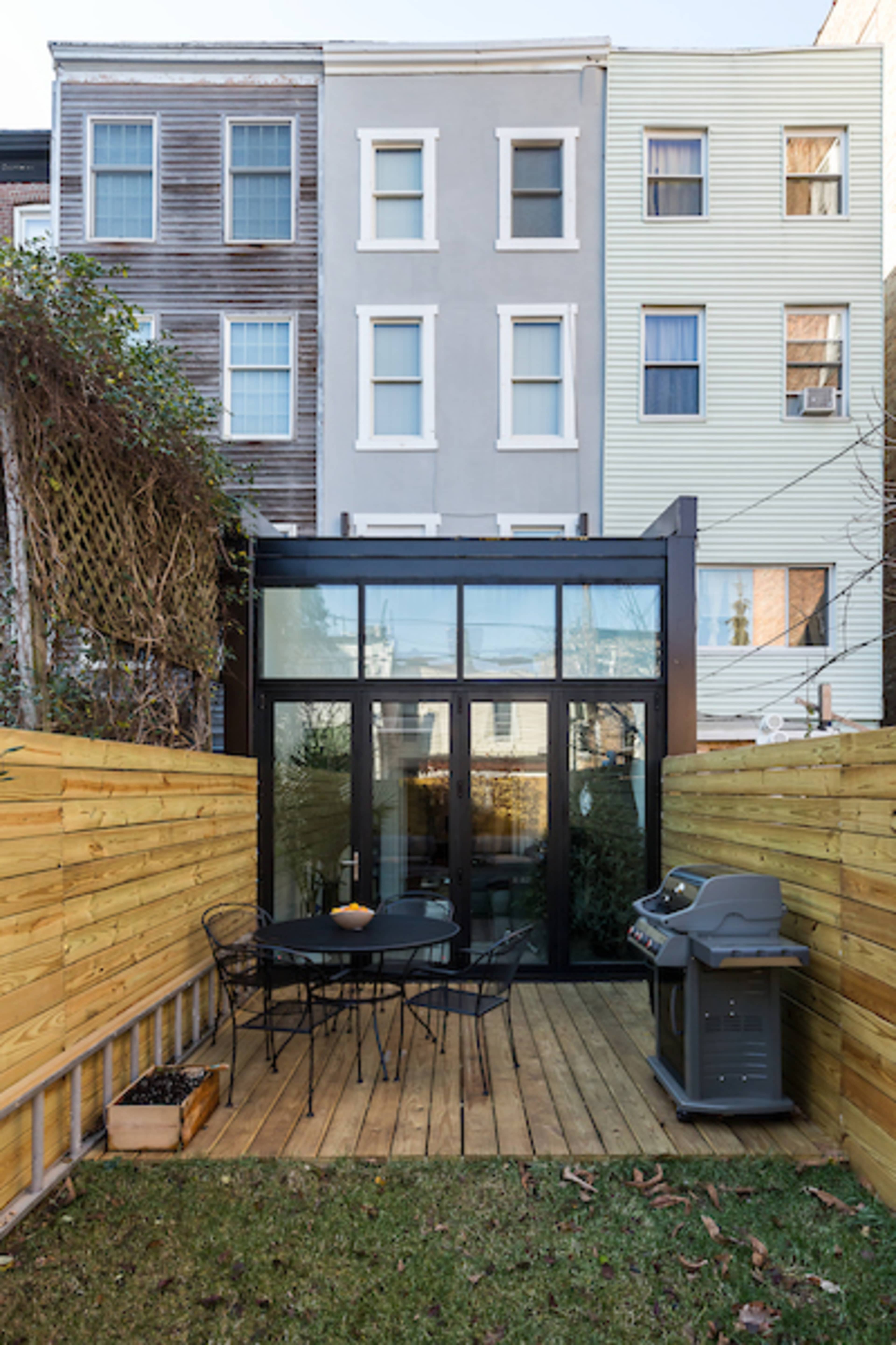 A small outdoor patio with wooden decking, black metal furniture, and a grill, surrounded by wooden fences and adjacent to multi-colored townhouses.